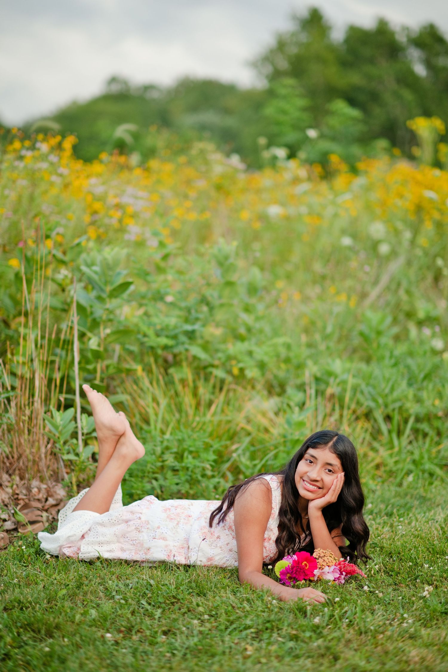 Young woman in floral dress lying on grass smiling, holding pink flowers with yellow wildflowers in background.