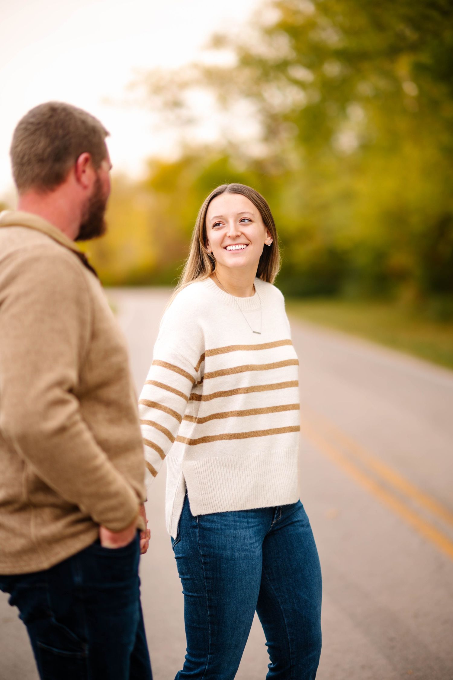 A couple in casual fall attire share playful moments along a tree-lined path during golden hour.