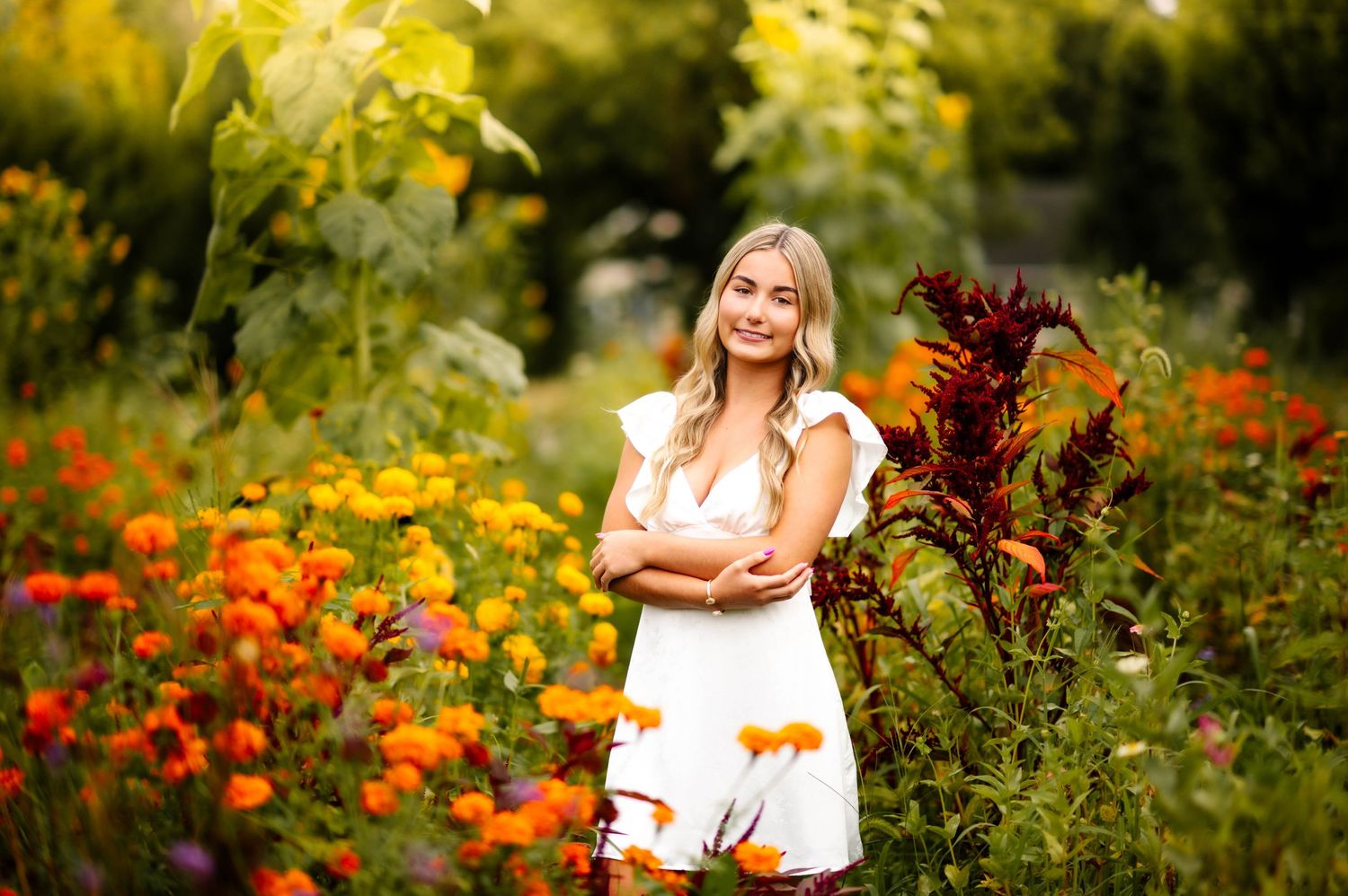 Young blonde woman in white dress smiling among vibrant orange and red flowers in a lush garden.