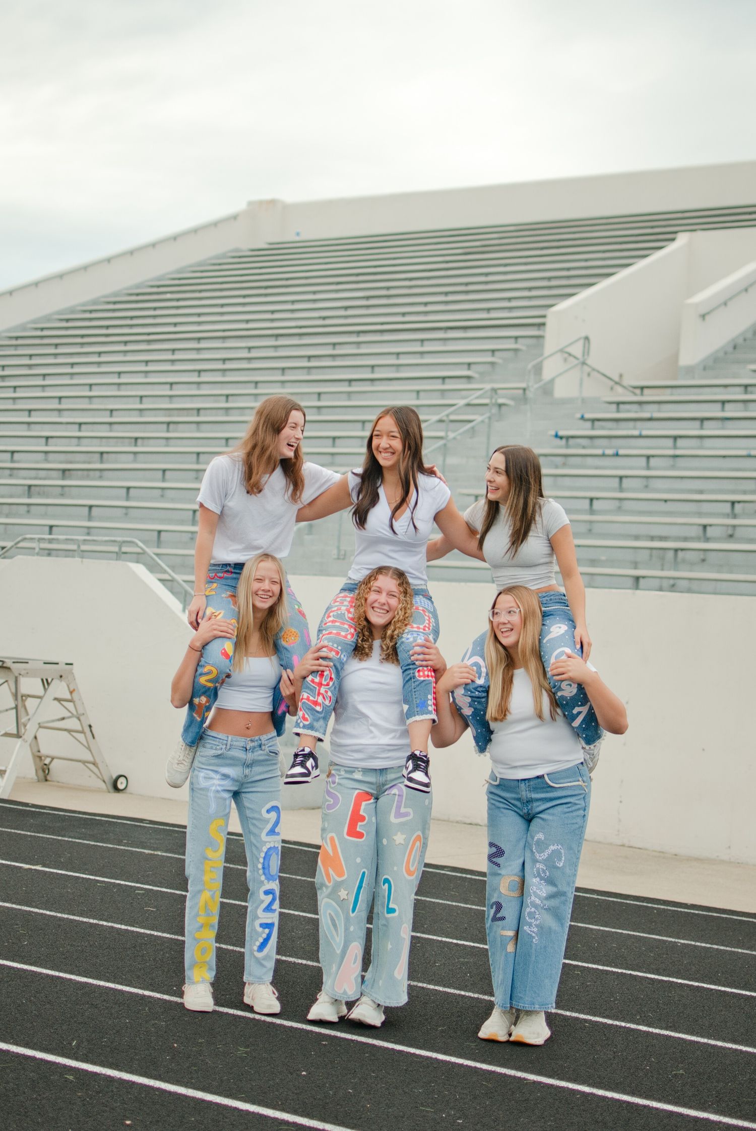 Six girls in white tops and painted senior jeans pose in a fun pyramid formation on a track.