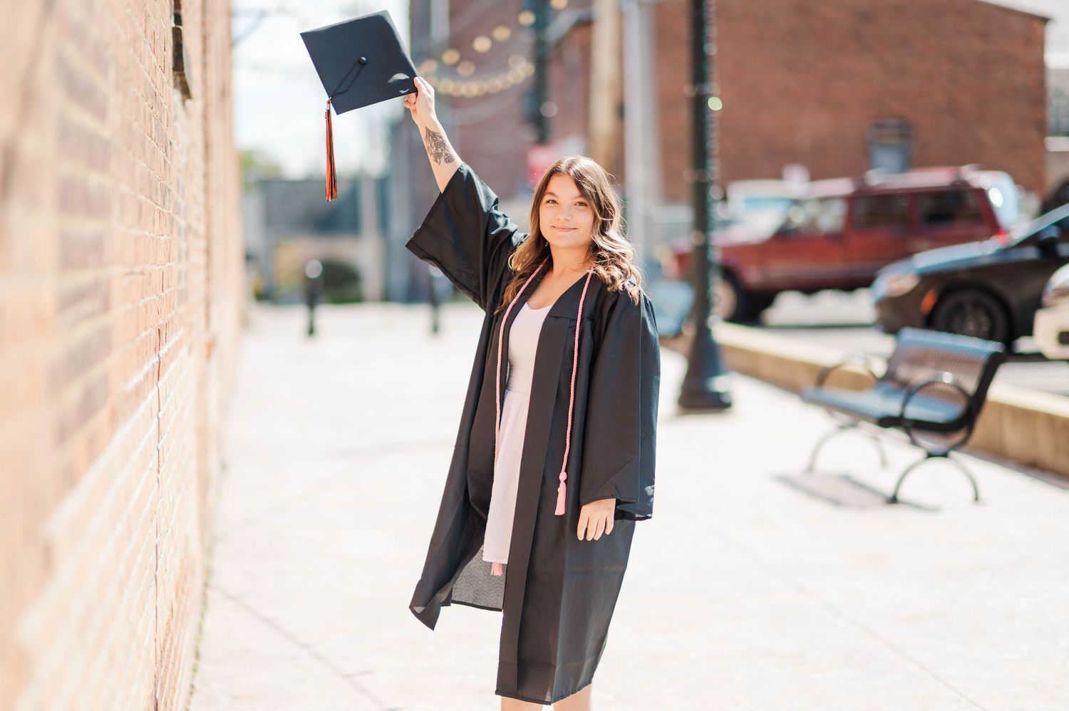 Female graduate in black cap and gown holding mortarboard up while posing on a city sidewalk.