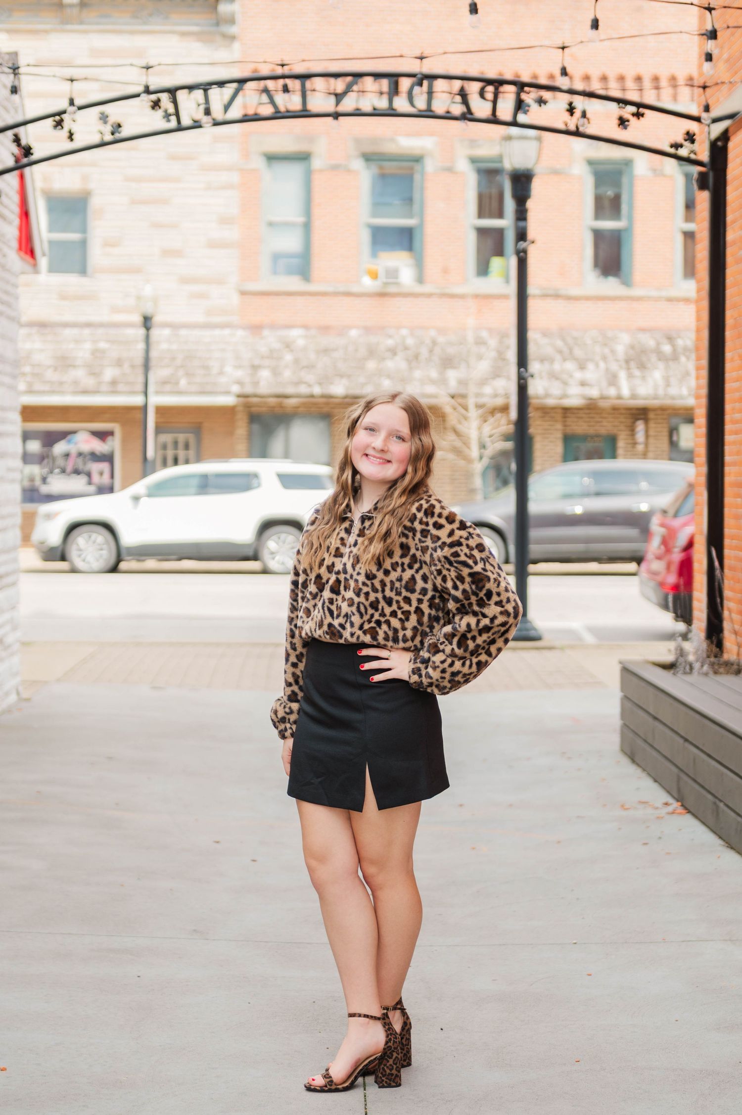 Young woman smiling in leopard print top and black mini skirt posing under decorative arch on urban sidewalk.