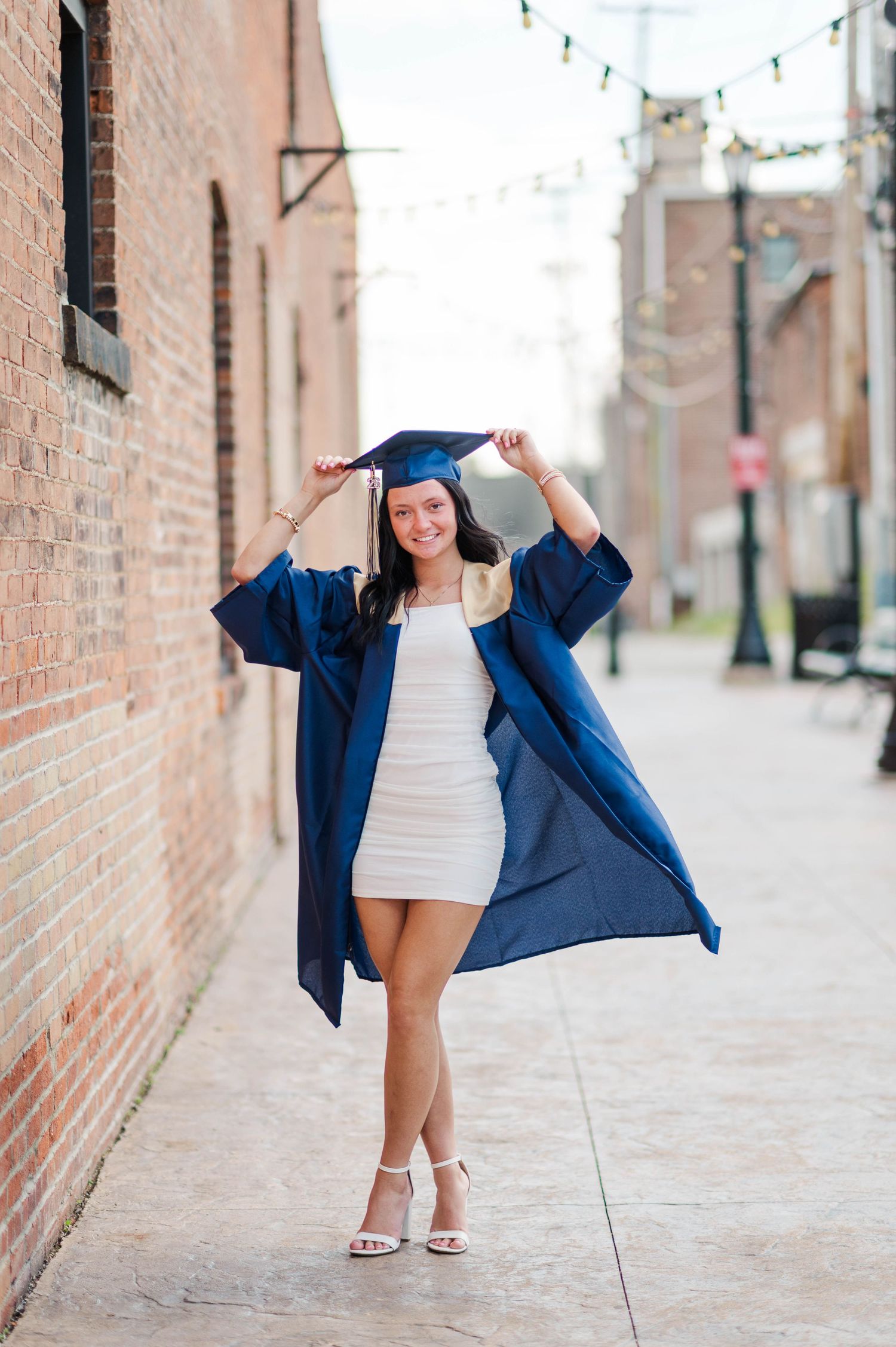 Smiling graduate poses in navy cap and gown over white dress in a sunny brick alley.