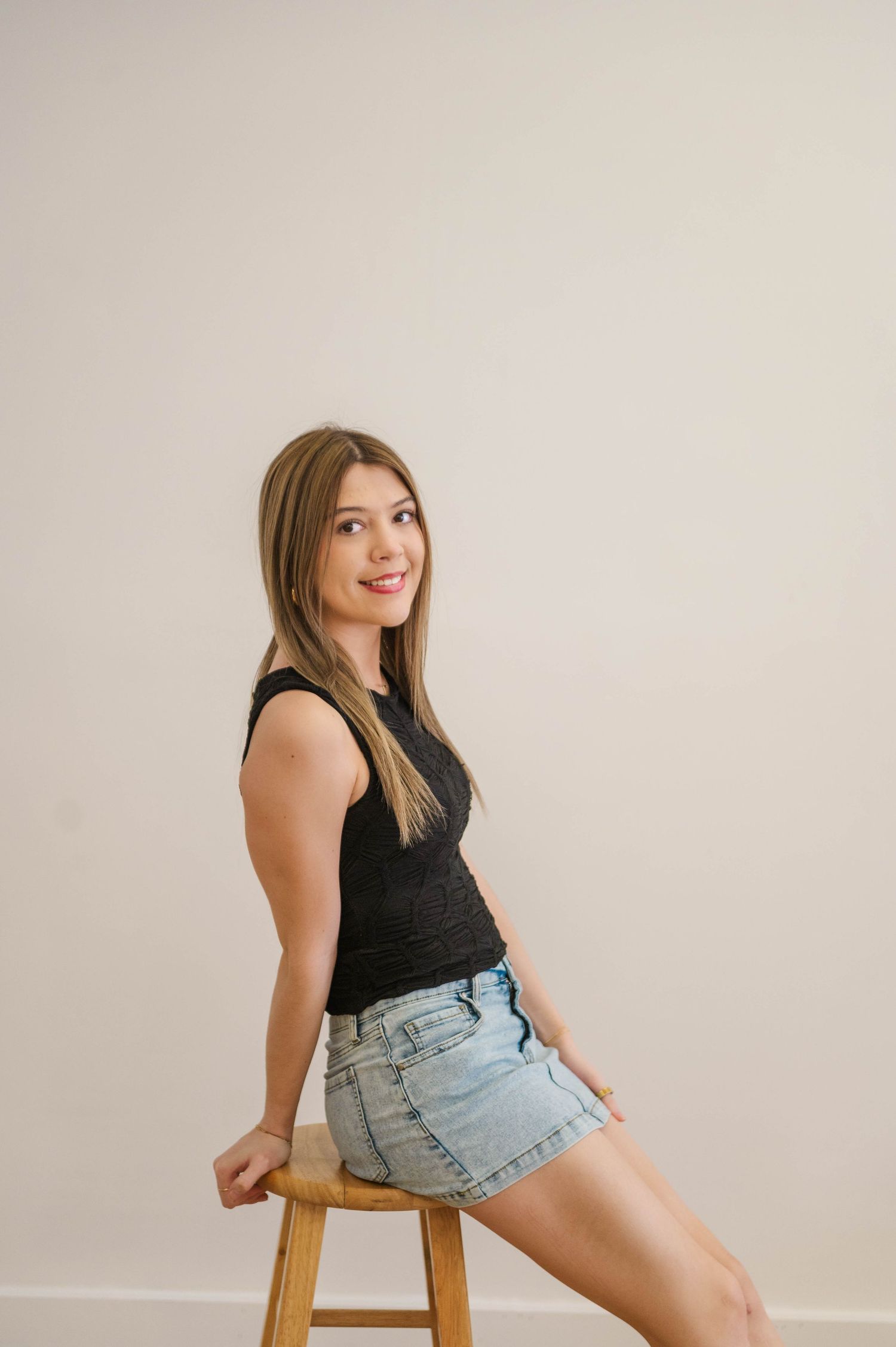 Smiling blonde woman in black sleeveless top and denim mini skirt sitting on wooden stool.