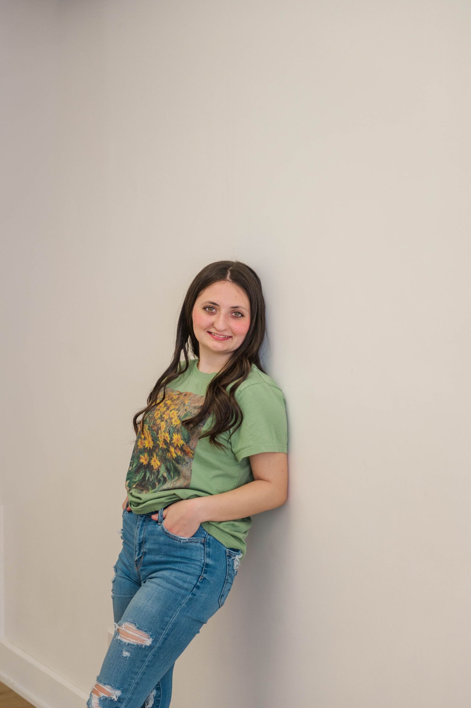 Young brunette woman leaning against white wall in green graphic tee and ripped jeans.