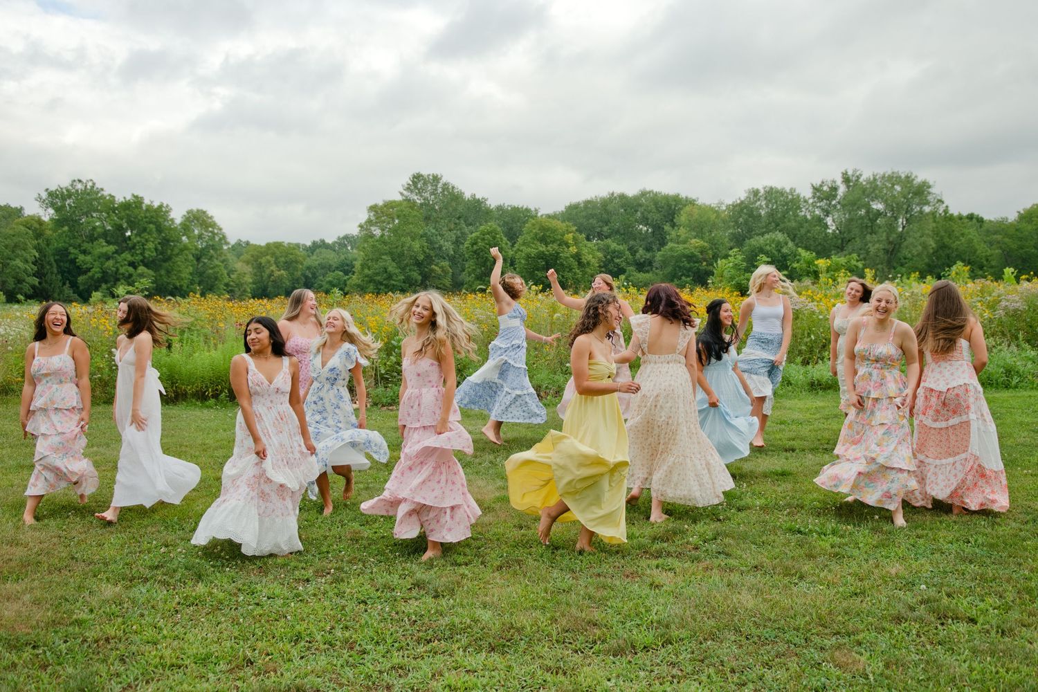 A group of women in colorful pastel dresses dancing joyfully in a lush green field on a cloudy day.