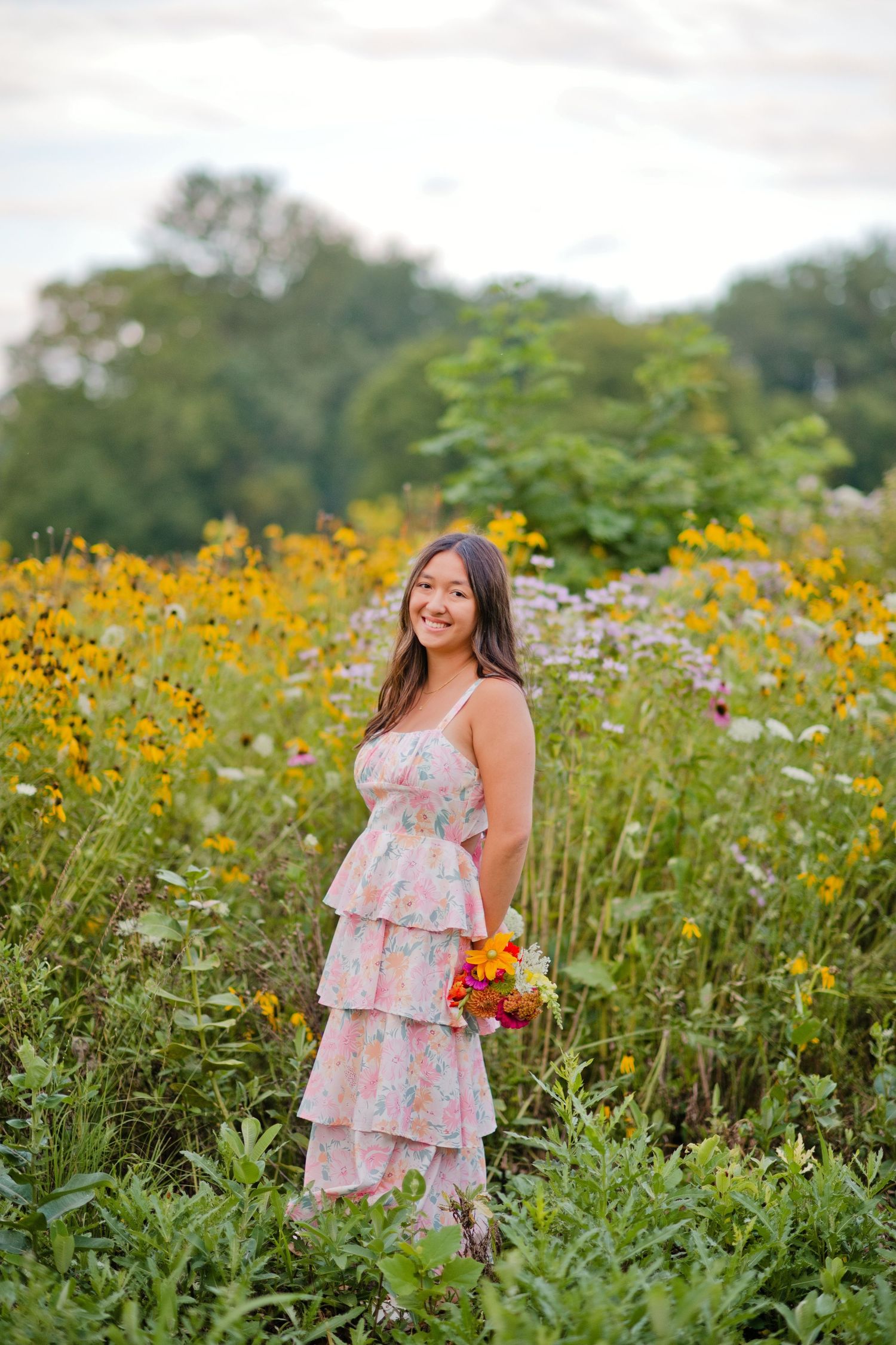 A smiling young woman in a pink floral tiered dress stands among yellow wildflowers holding a colorful bouquet.