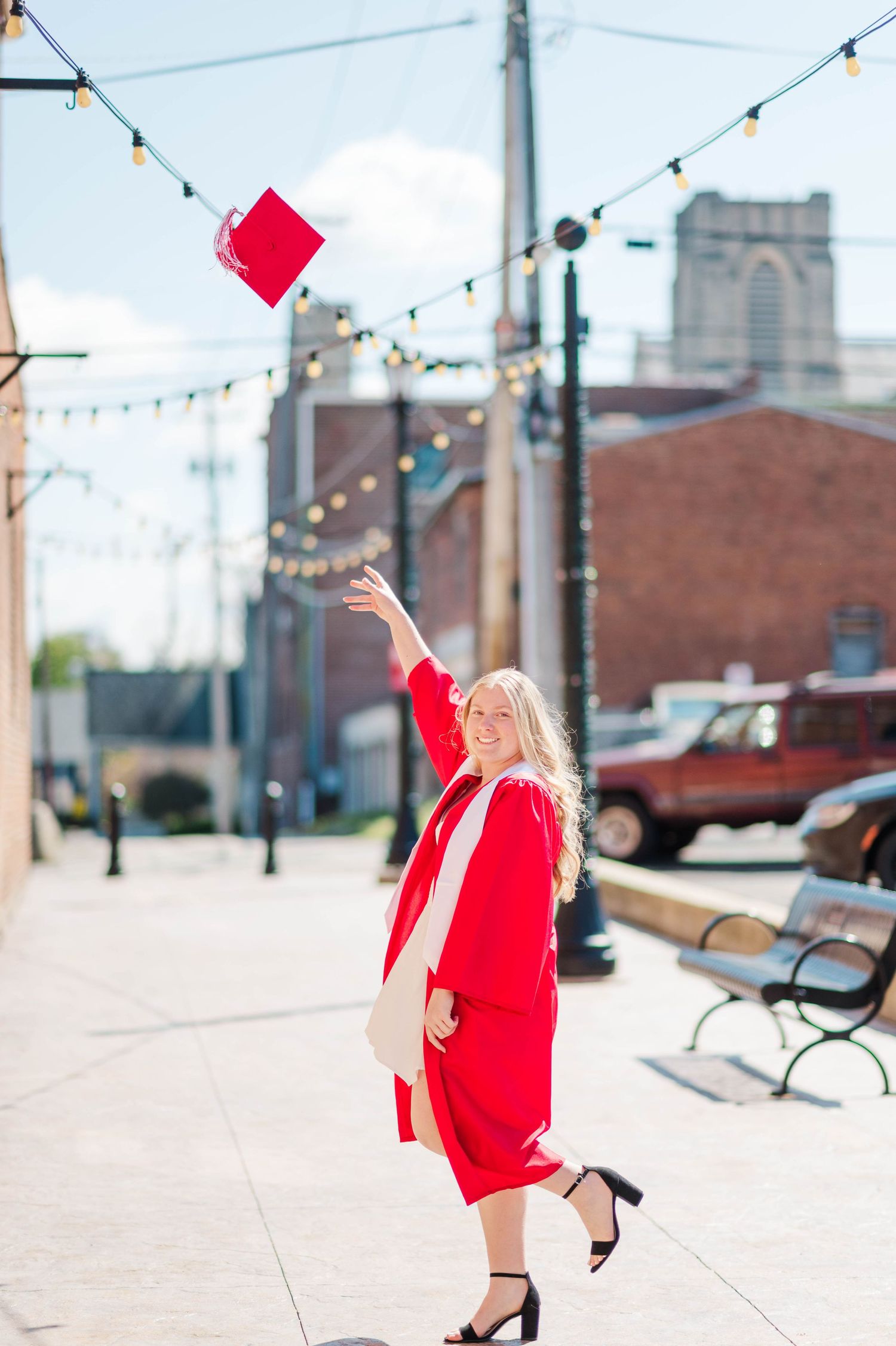 Graduate in red cap and gown tossing mortarboard into air on sunny small-town street with string lights.