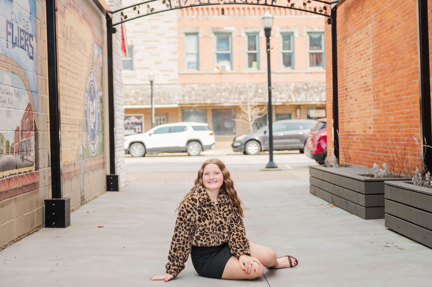 Young woman in leopard print jacket sits cross-legged on ground in outdoor urban corridor with brick walls.