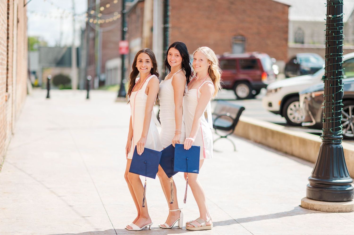 Three young women in matching white tops and blue shorts posing together on sunny sidewalk.