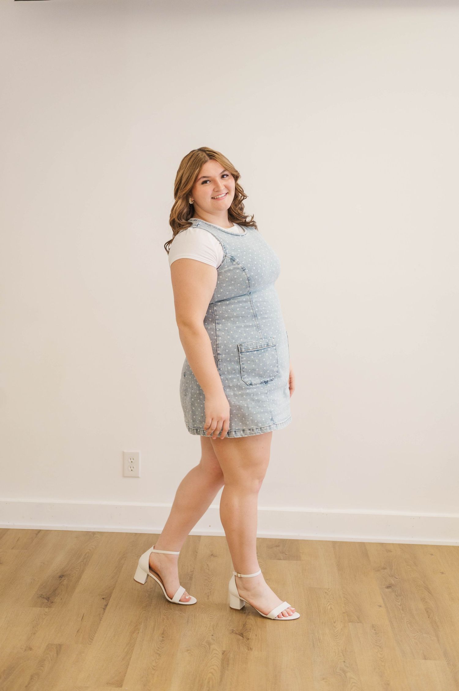 Young woman in denim jumper dress and white heels posing on hardwood floor against white wall.