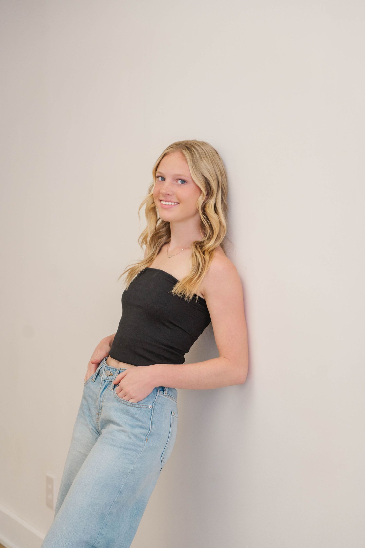 Blonde teen girl leans against white wall smiling, wearing black strapless top and light wash jeans.