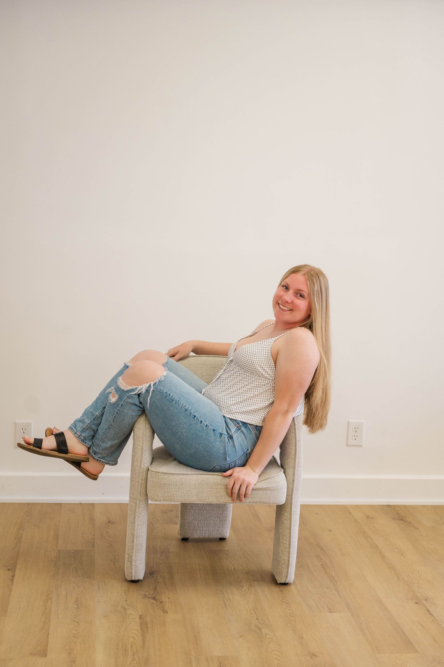 Smiling blonde woman in white tank and ripped jeans relaxing in modern chair on hardwood floor.