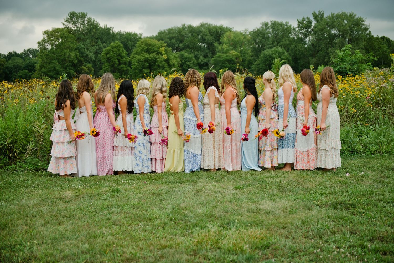 A row of bridesmaids in colorful pastel dresses holding bouquets stand together in a lush green field.