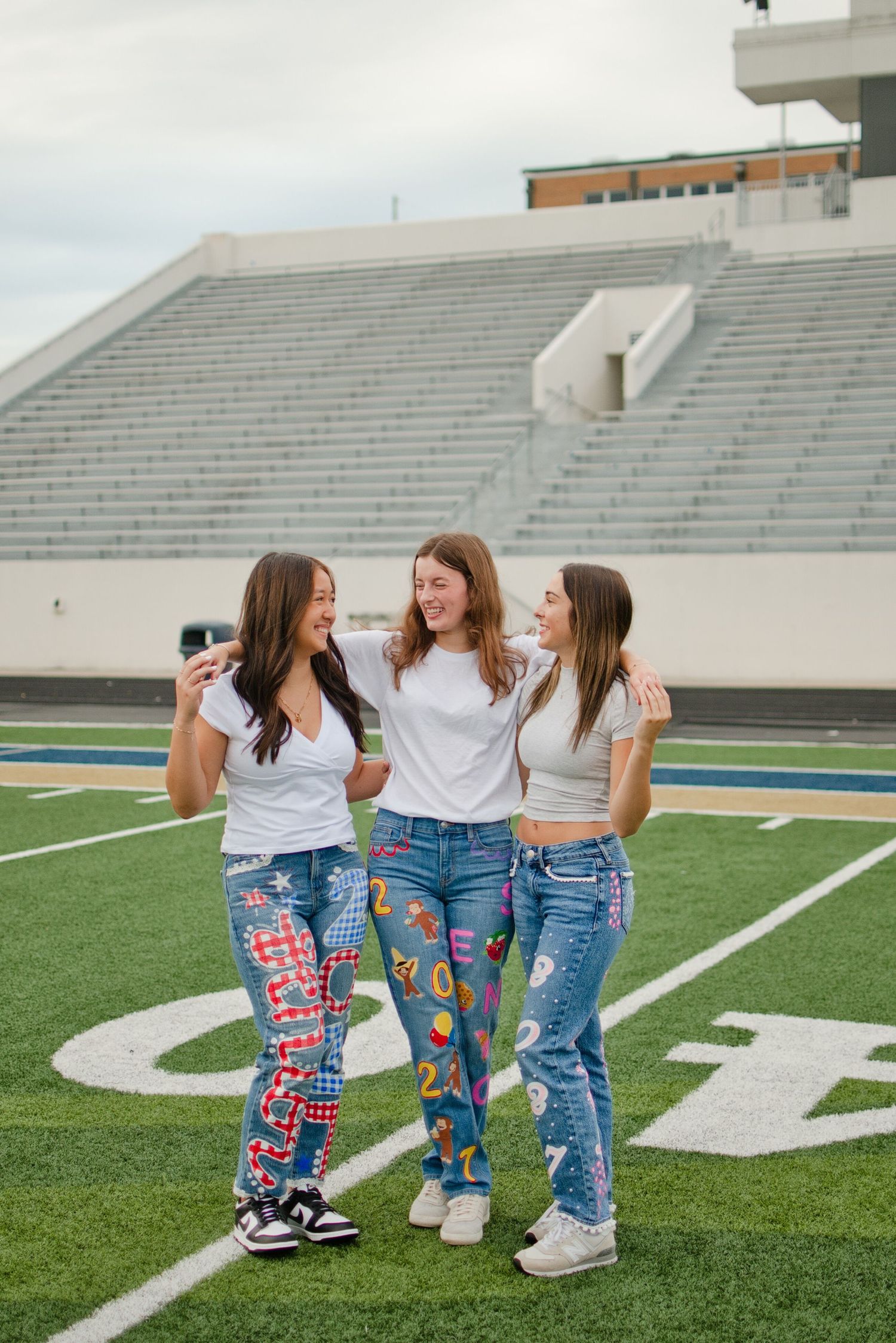 Three teen girls in white tees and custom painted jeans laugh together on a football field.