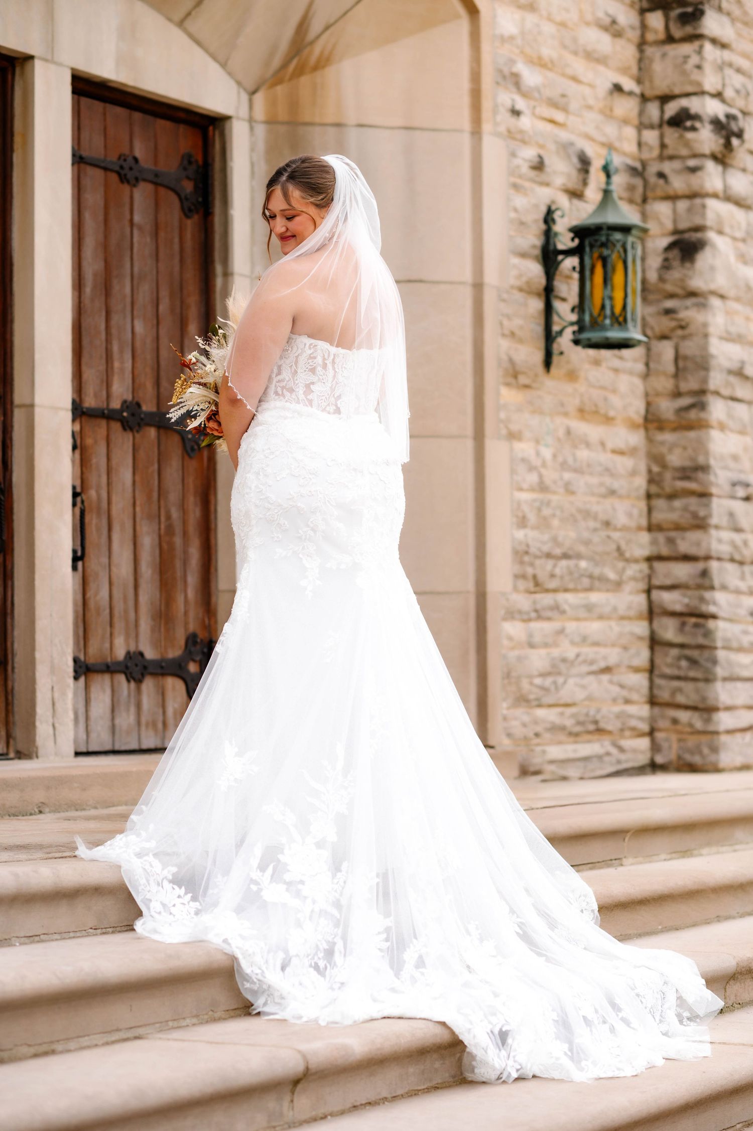 Bride in lace wedding gown poses along stone church exterior wall.