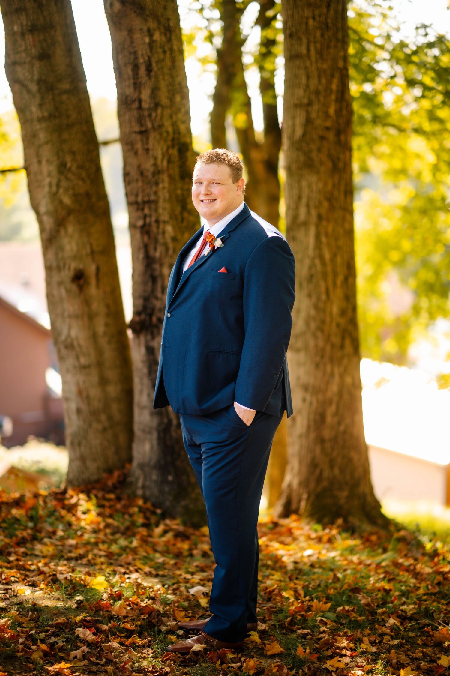 Groom in navy suit with red tie poses among fall leaves and trees.
