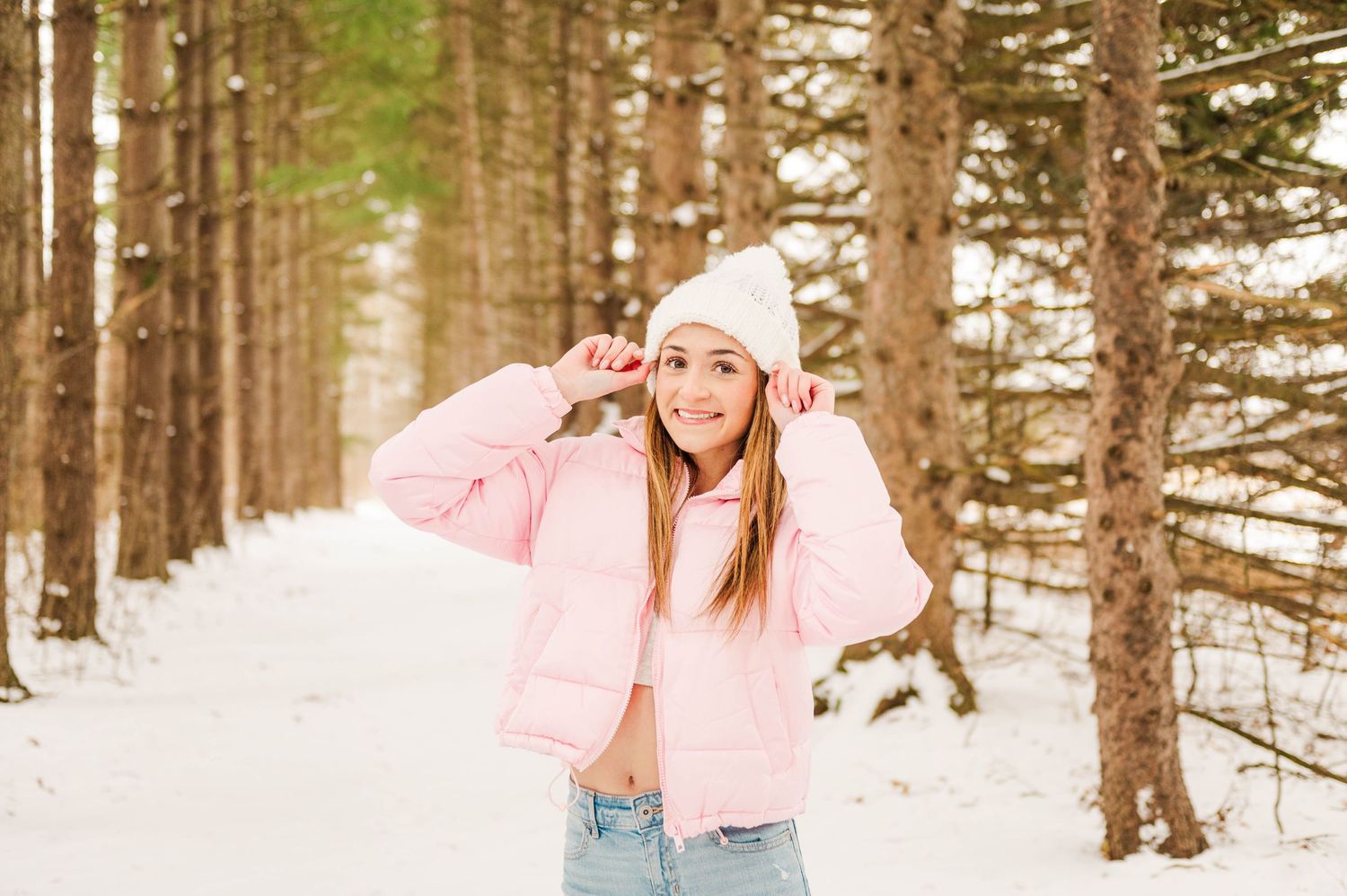 Winter photoshoot in a snowy forest with someone wearing a pink jacket and white beanie.
