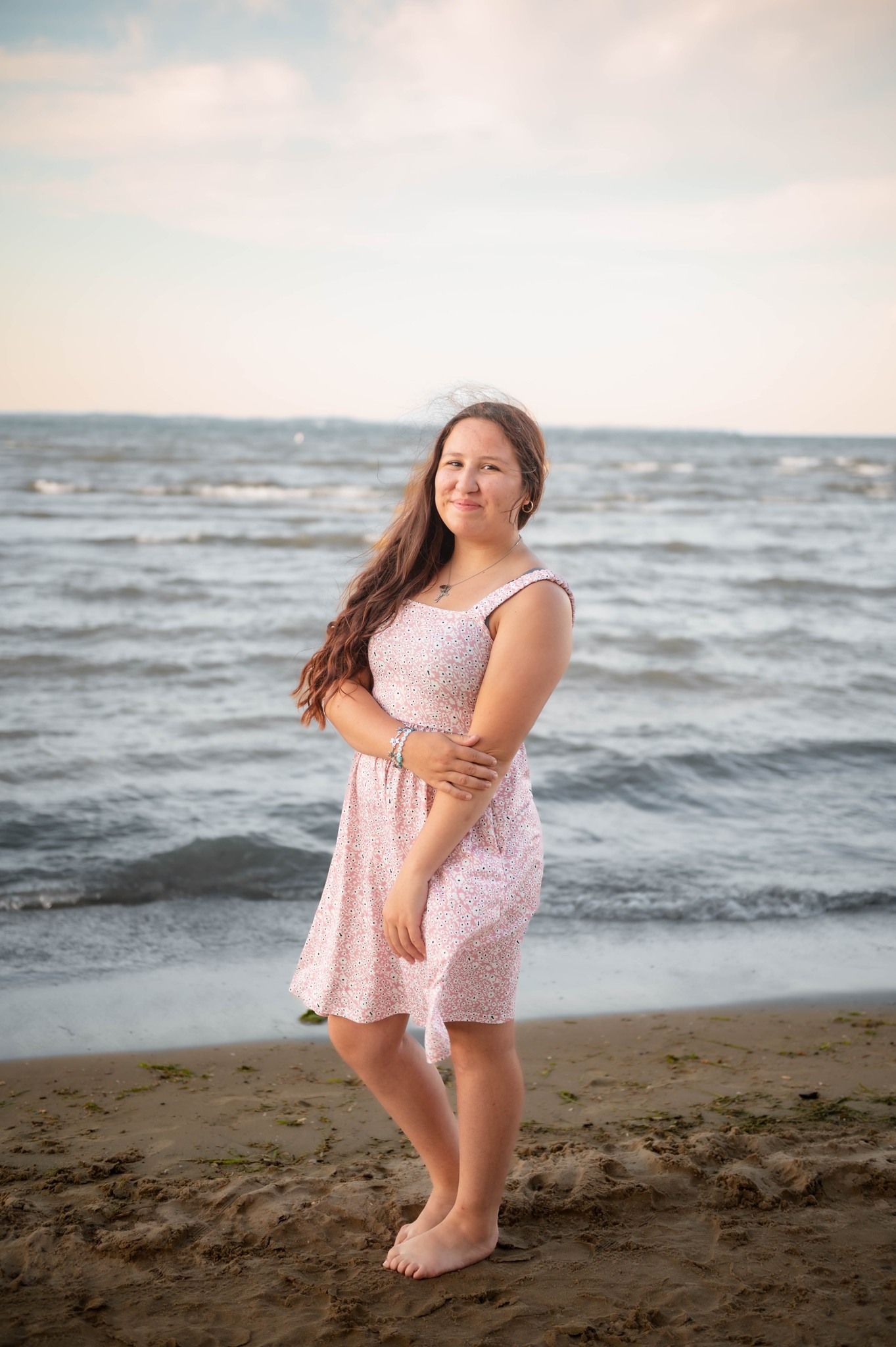 A person in a light pink dress dances playfully on the beach with waves in the background.
