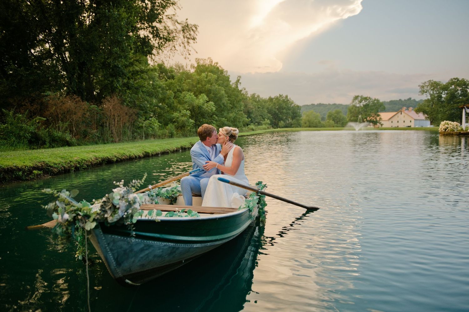 Newlywed couple sharing a romantic moment in a flower-adorned rowboat on a calm lake at sunset.