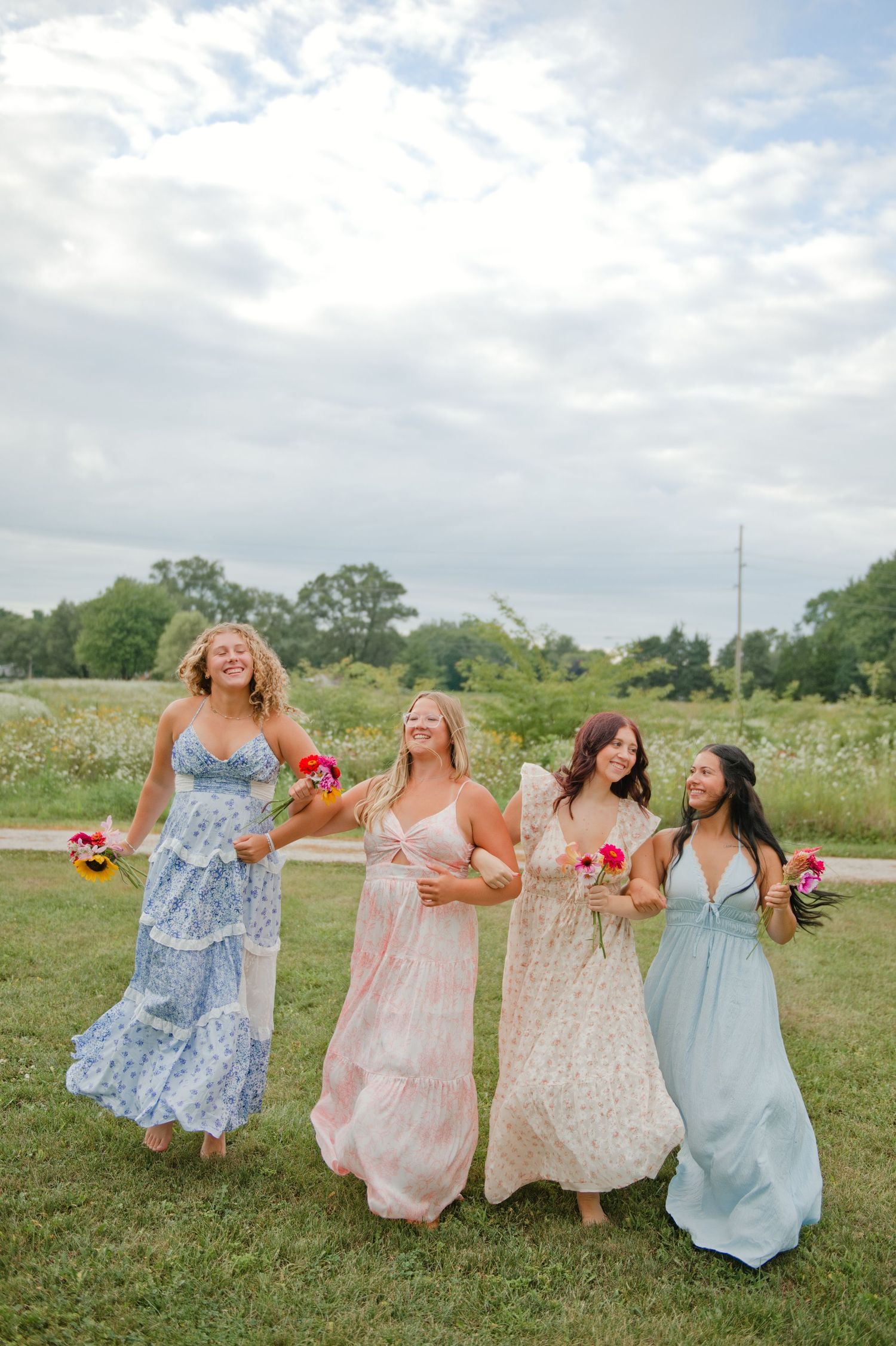 Four women in floral maxi dresses laughing and walking together in a meadow.