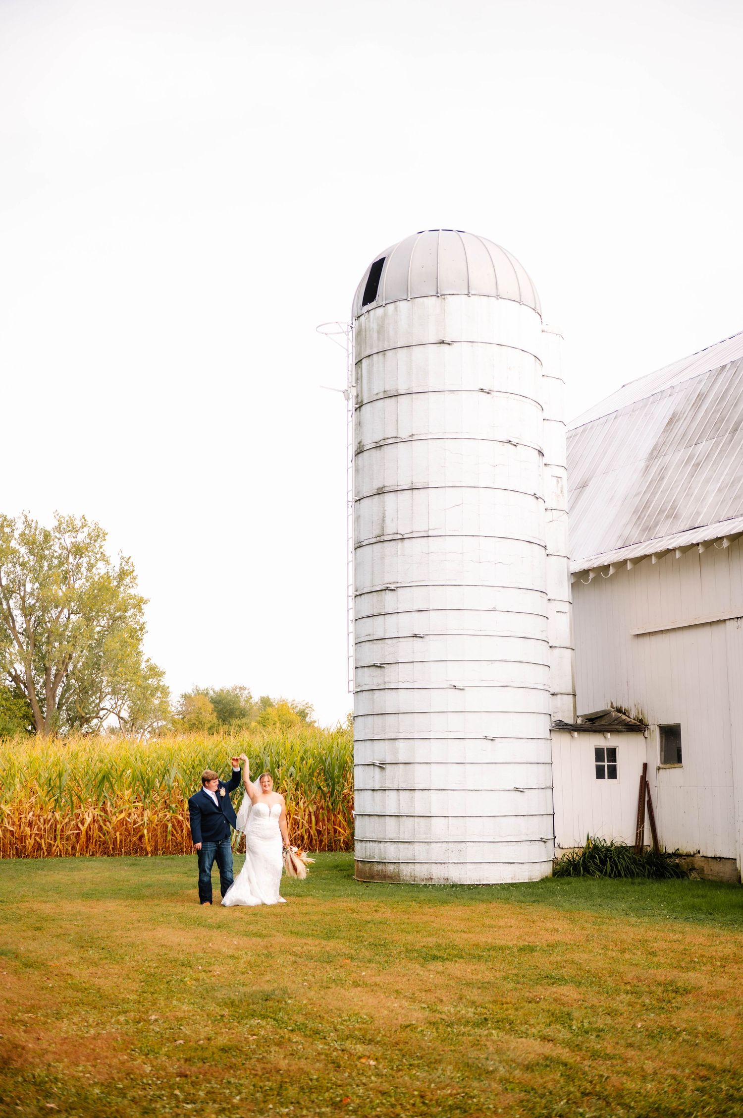 Wedding couple poses for photos near white farm silos and cornfields at sunset.