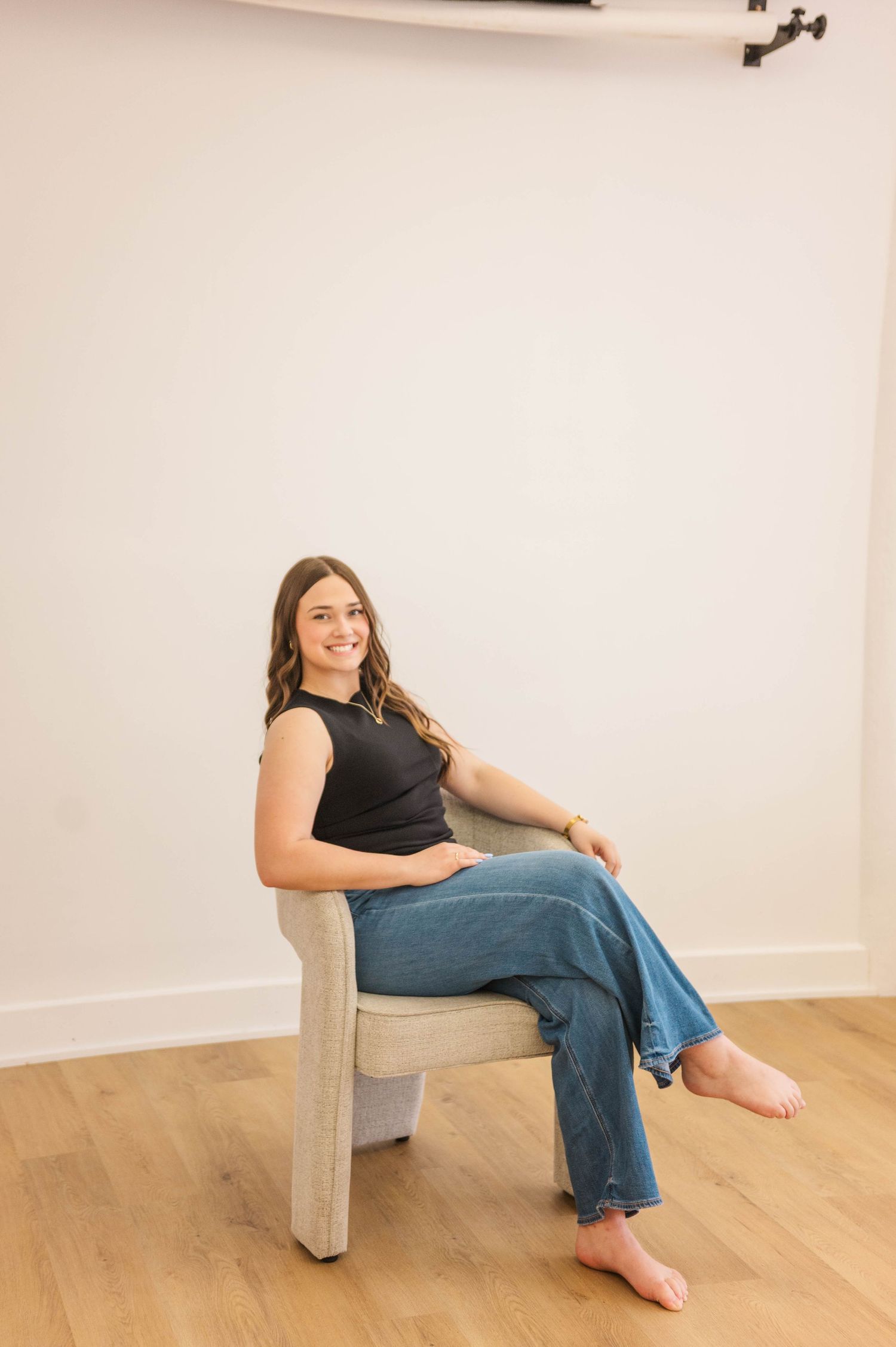 Young woman in black tank top and jeans sitting relaxed in a modern chair on hardwood floor.