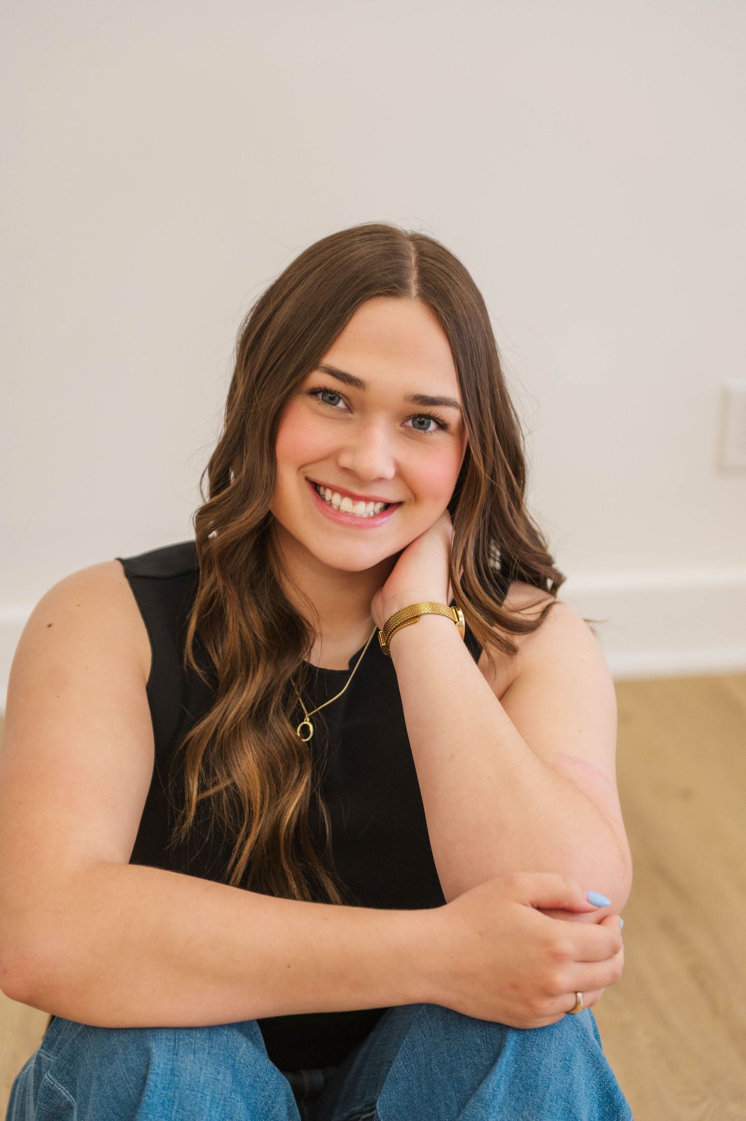Teenage girl with wavy brown hair sits on floor in black top and jeans, smiling with hand on chin.