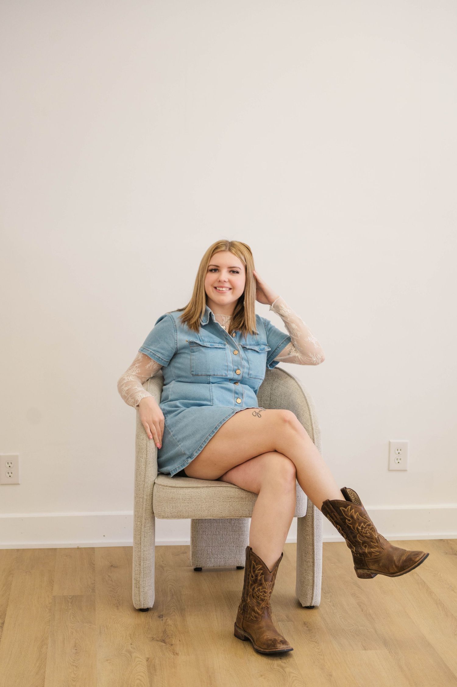 Blonde woman in denim dress and cowboy boots sitting cross-legged in a modern chair on hardwood floor.
