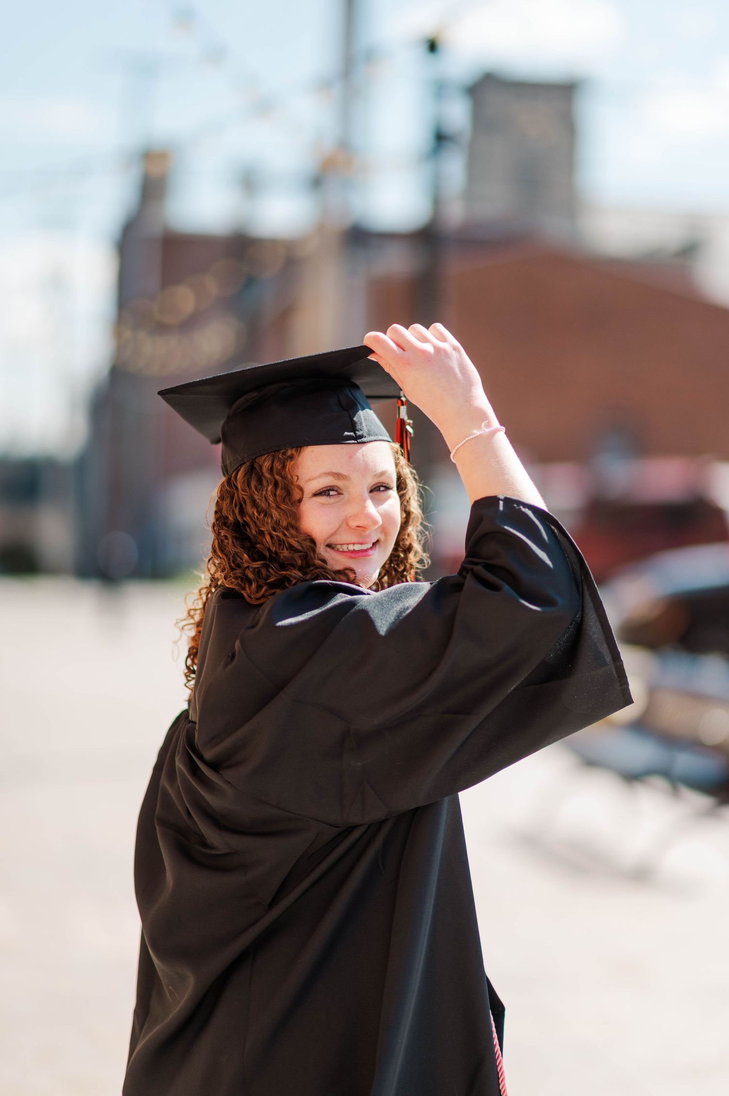 Curly-haired graduate in black cap and gown smiles over shoulder on bright sunny urban street.