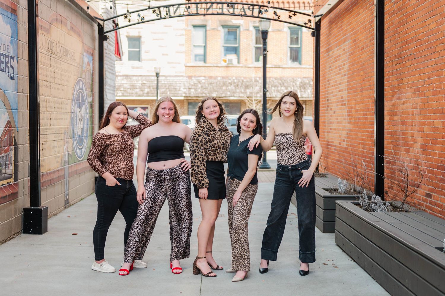 Five young women in coordinated leopard and black outfits smiling together in a downtown alley with brick buildings.