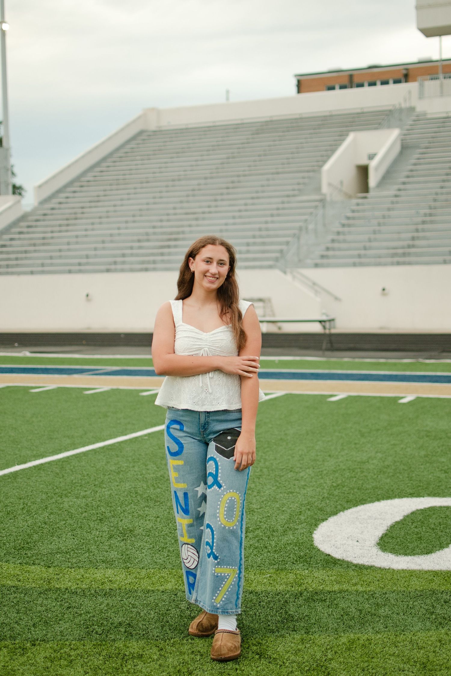 Young woman smiles on football field wearing white top and colorful painted senior jeans.
