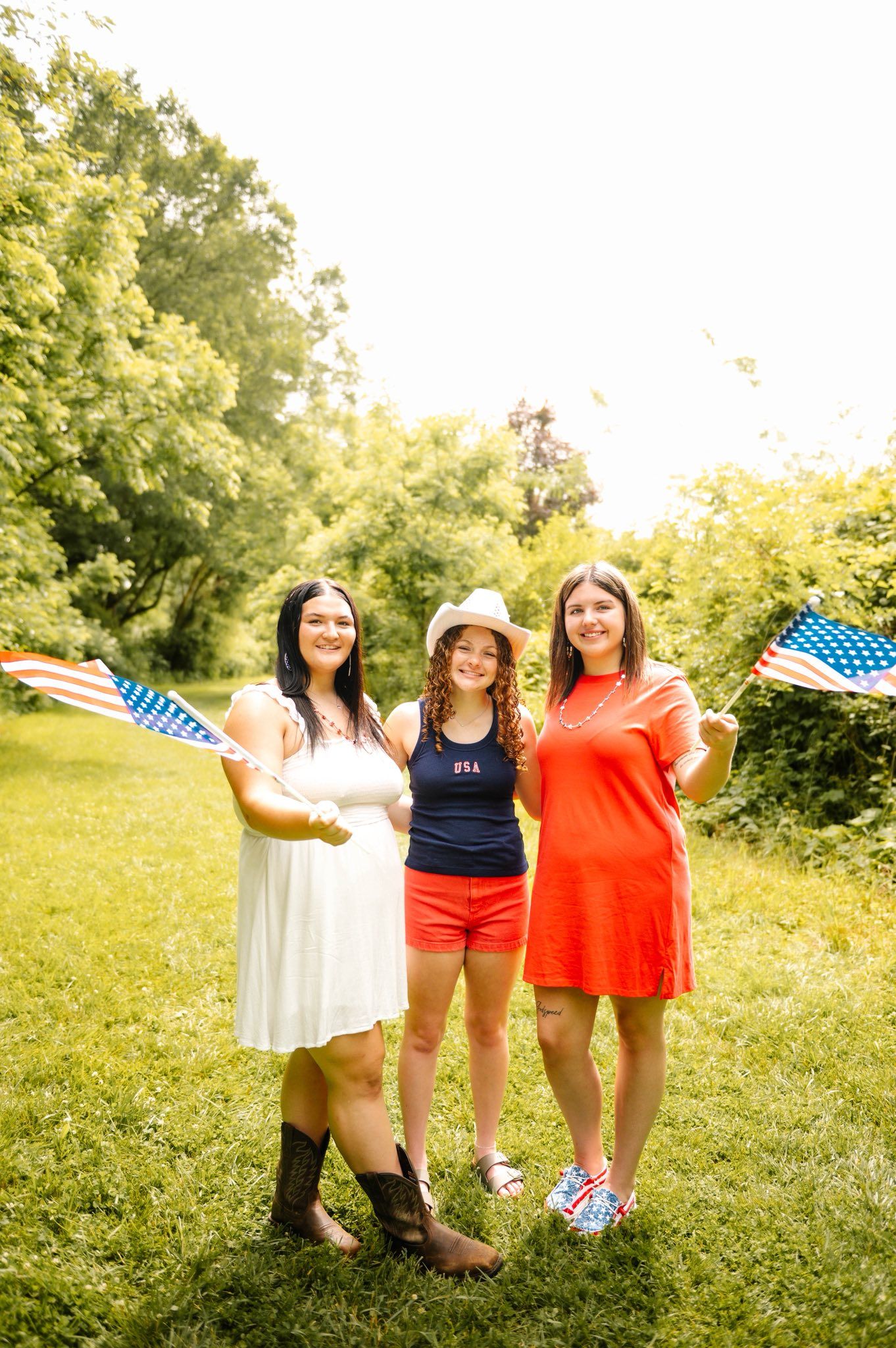 Three friends in patriotic colors wave American flags while posing outdoors.