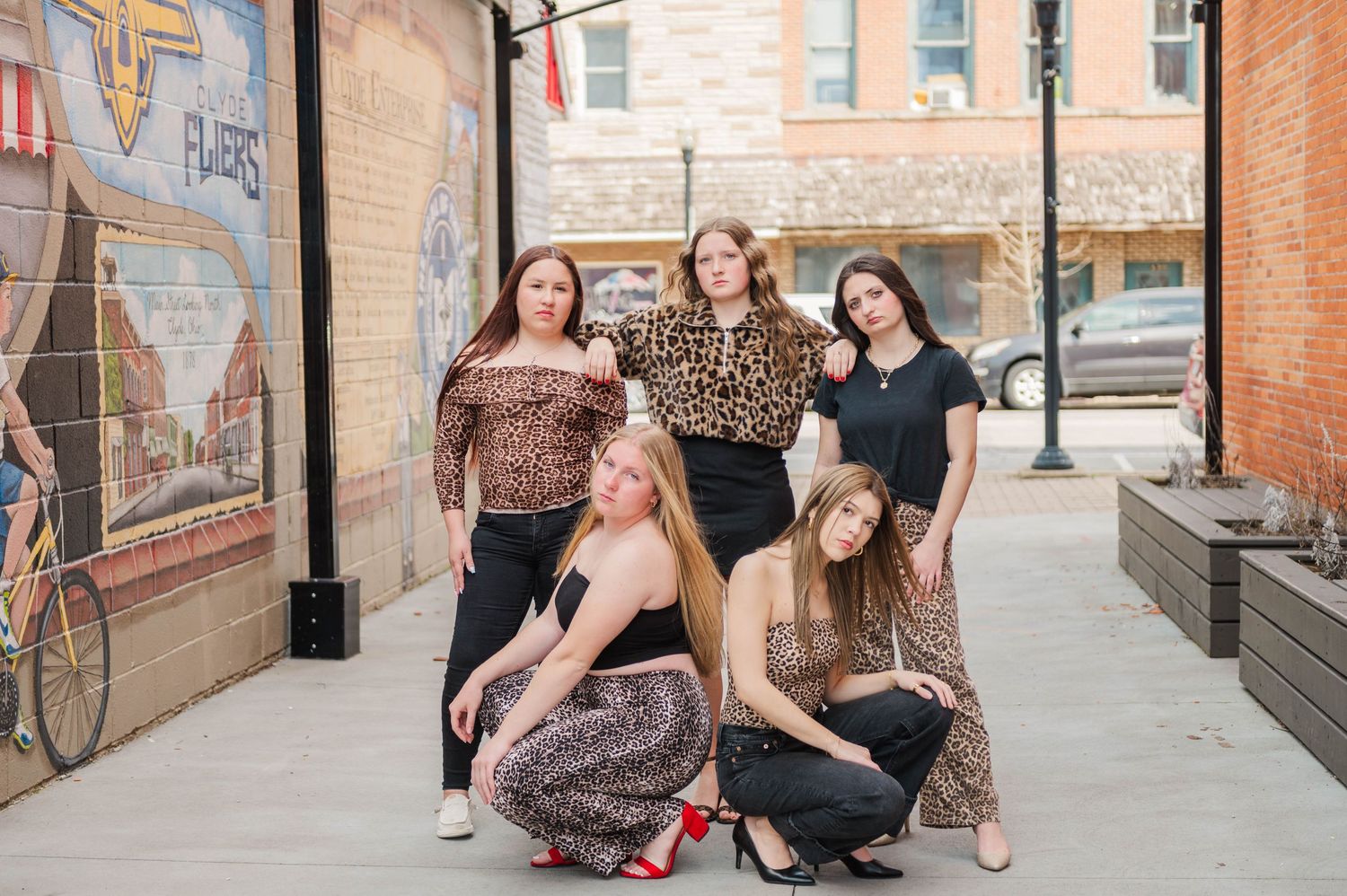 Five young women posing together on an urban sidewalk beside a colorful brick wall mural.