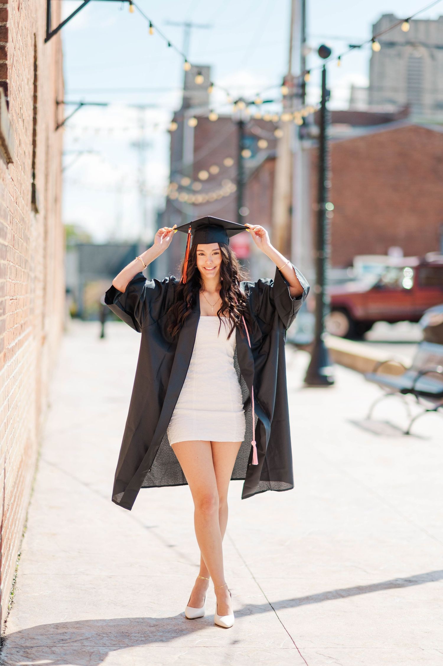 Smiling graduate in black cap and gown walks confidently along sunny urban brick alley sidewalk.