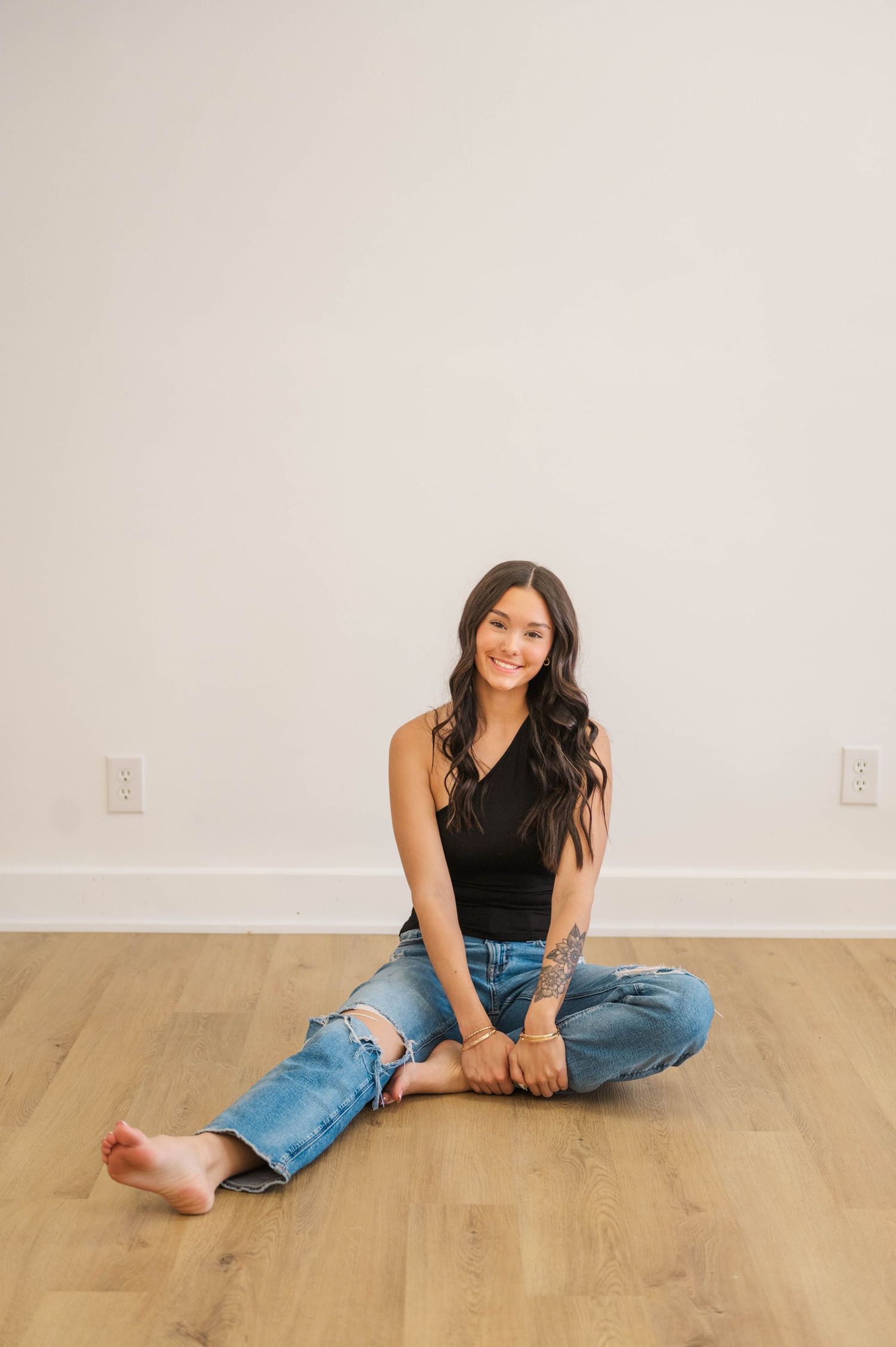 Woman in black tank and ripped jeans sitting cross-legged on hardwood floor, smiling.