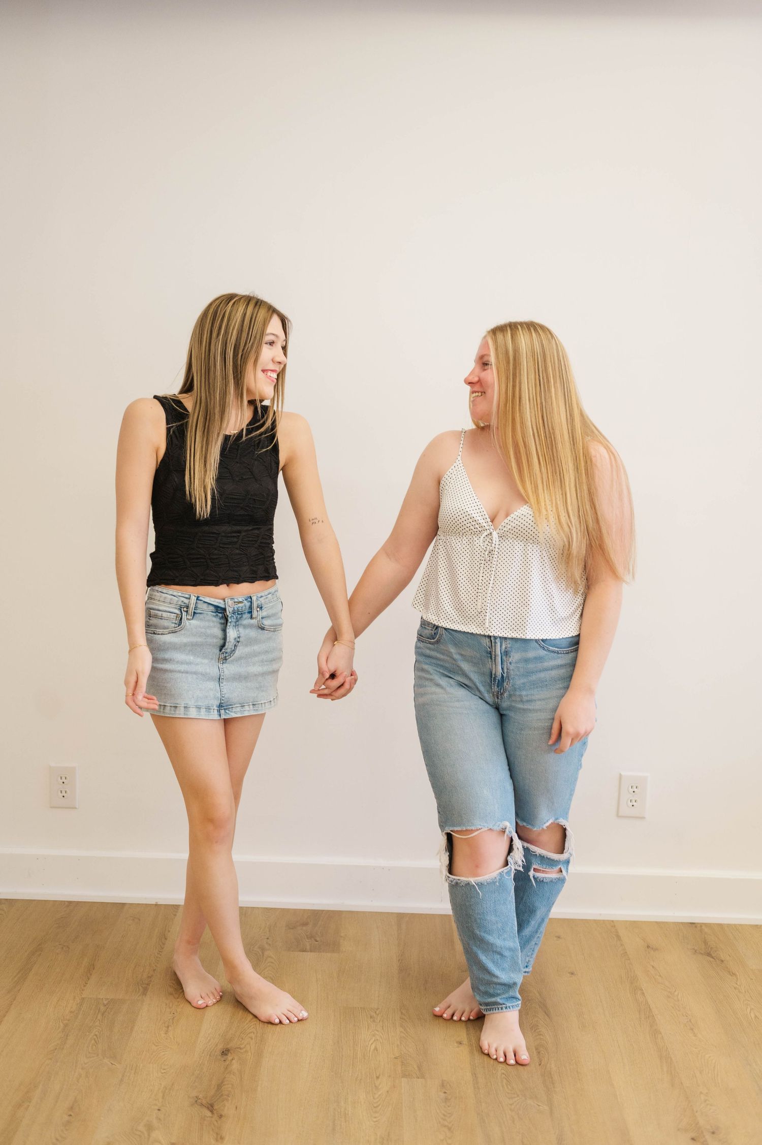 Two blonde women in casual outfits holding hands and laughing in a bright white room.