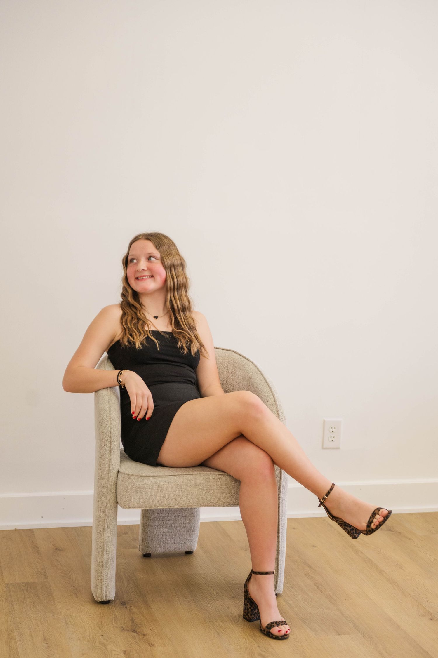 Young woman in black mini dress and heels seated in modern chair gazing upward in bright studio.