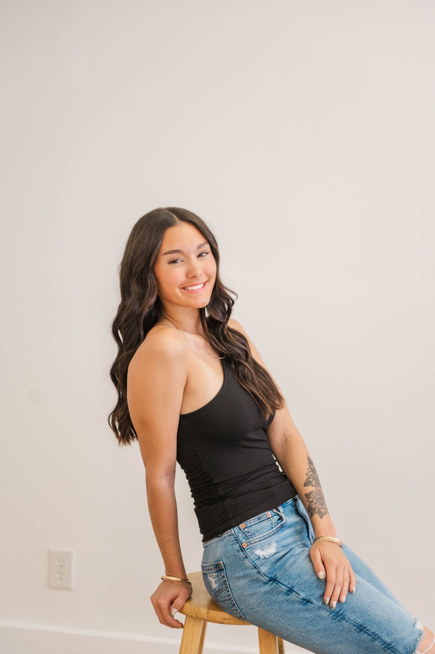 Young woman with long dark hair and tattoo sits on stool smiling, wearing black tank top and light blue jeans.