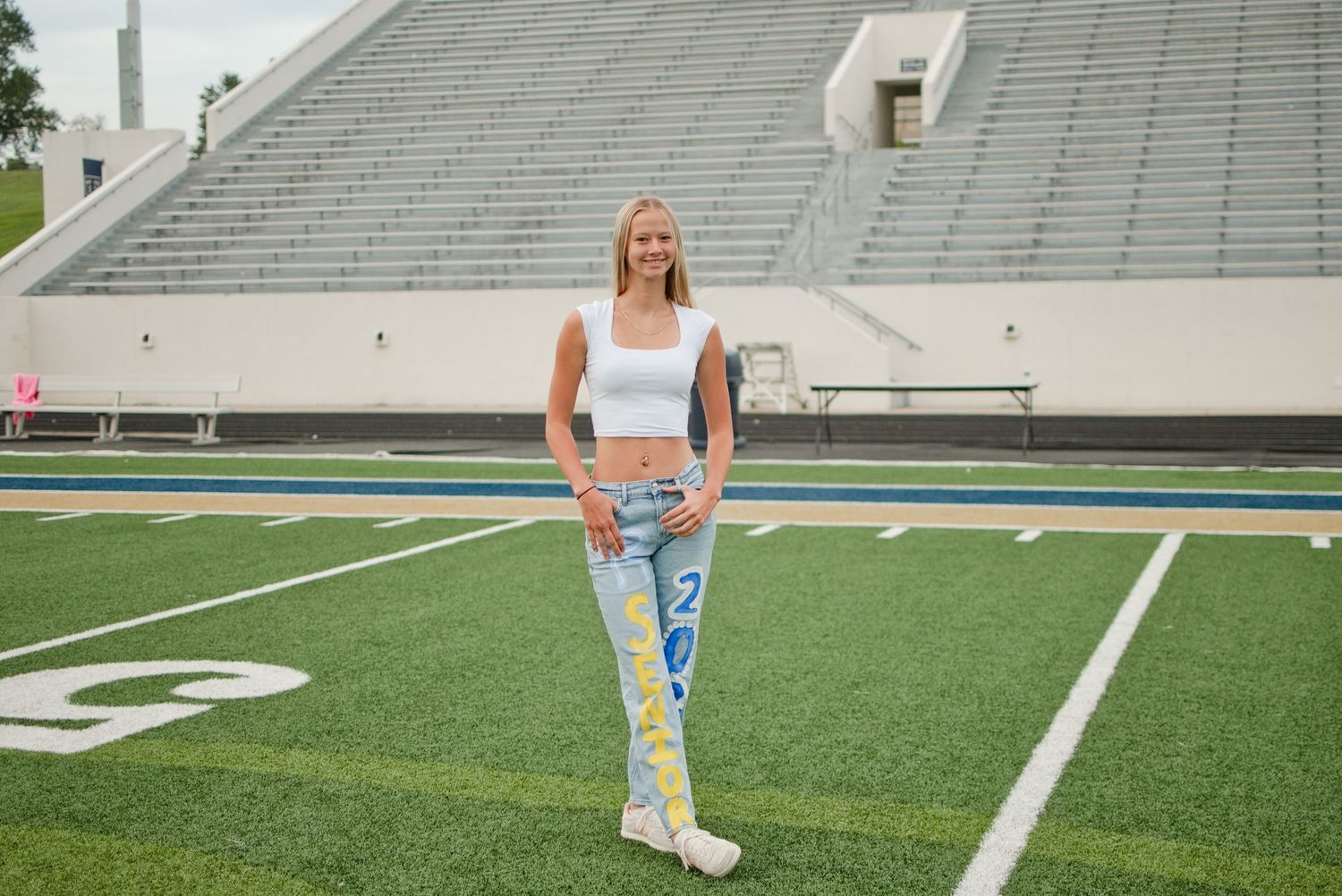 Blonde teenage girl in white crop top and decorated senior jeans poses confidently on a green football field.
