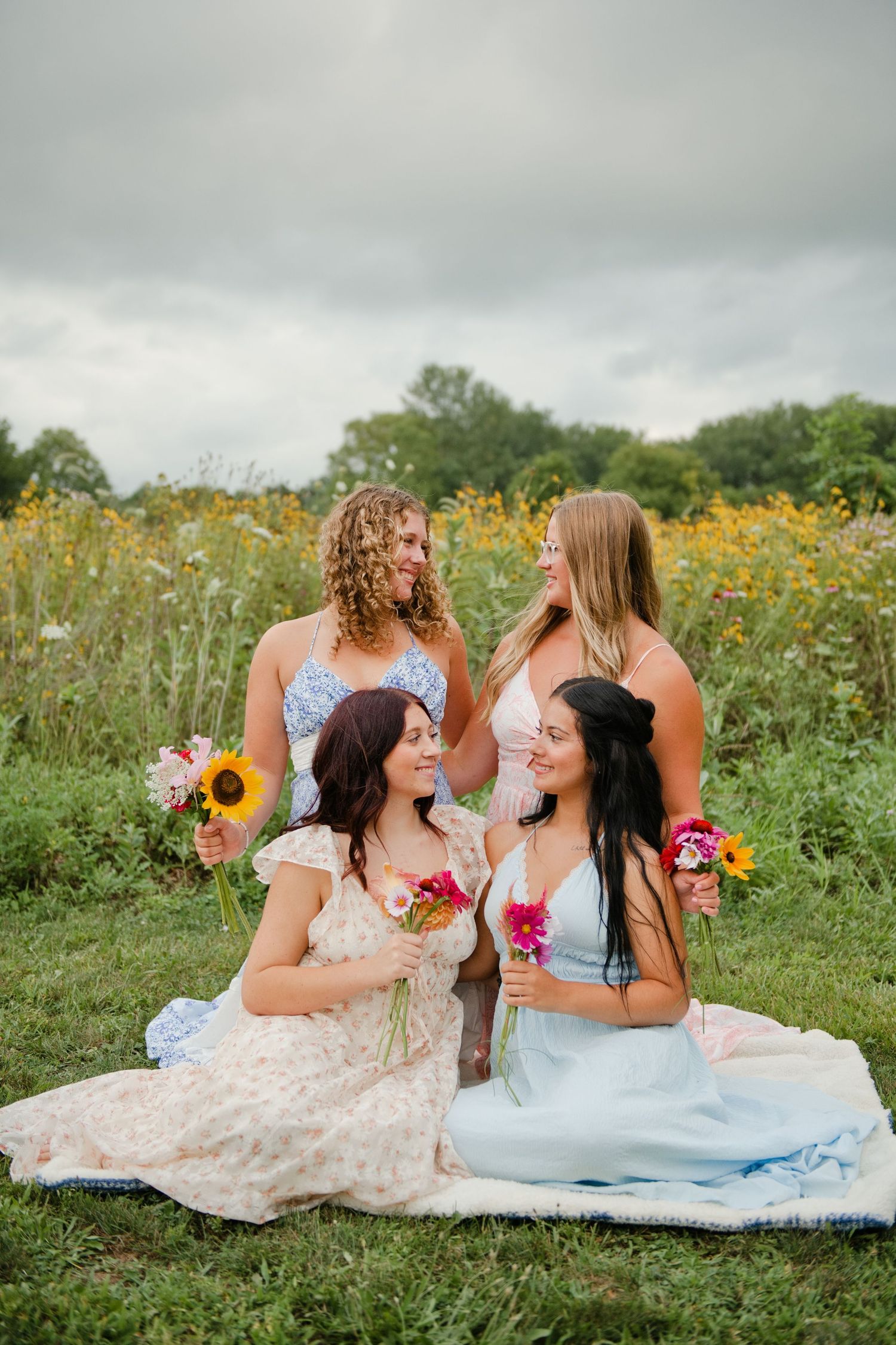 Four bridesmaids in pastel dresses sitting in a wildflower field holding colorful blooms.