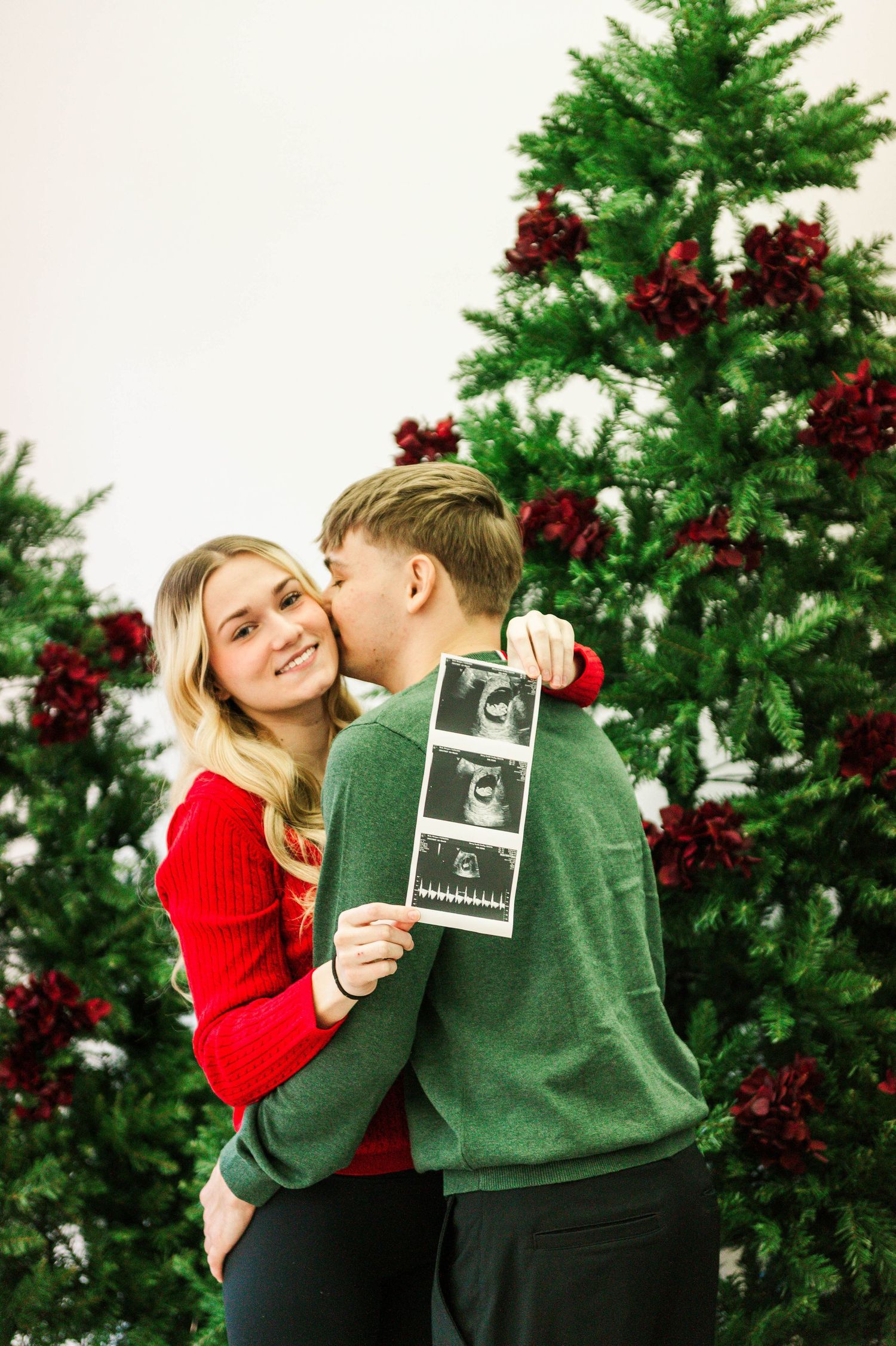 A couple shares a tender moment while holding ultrasound photos in front of a decorated Christmas tree.