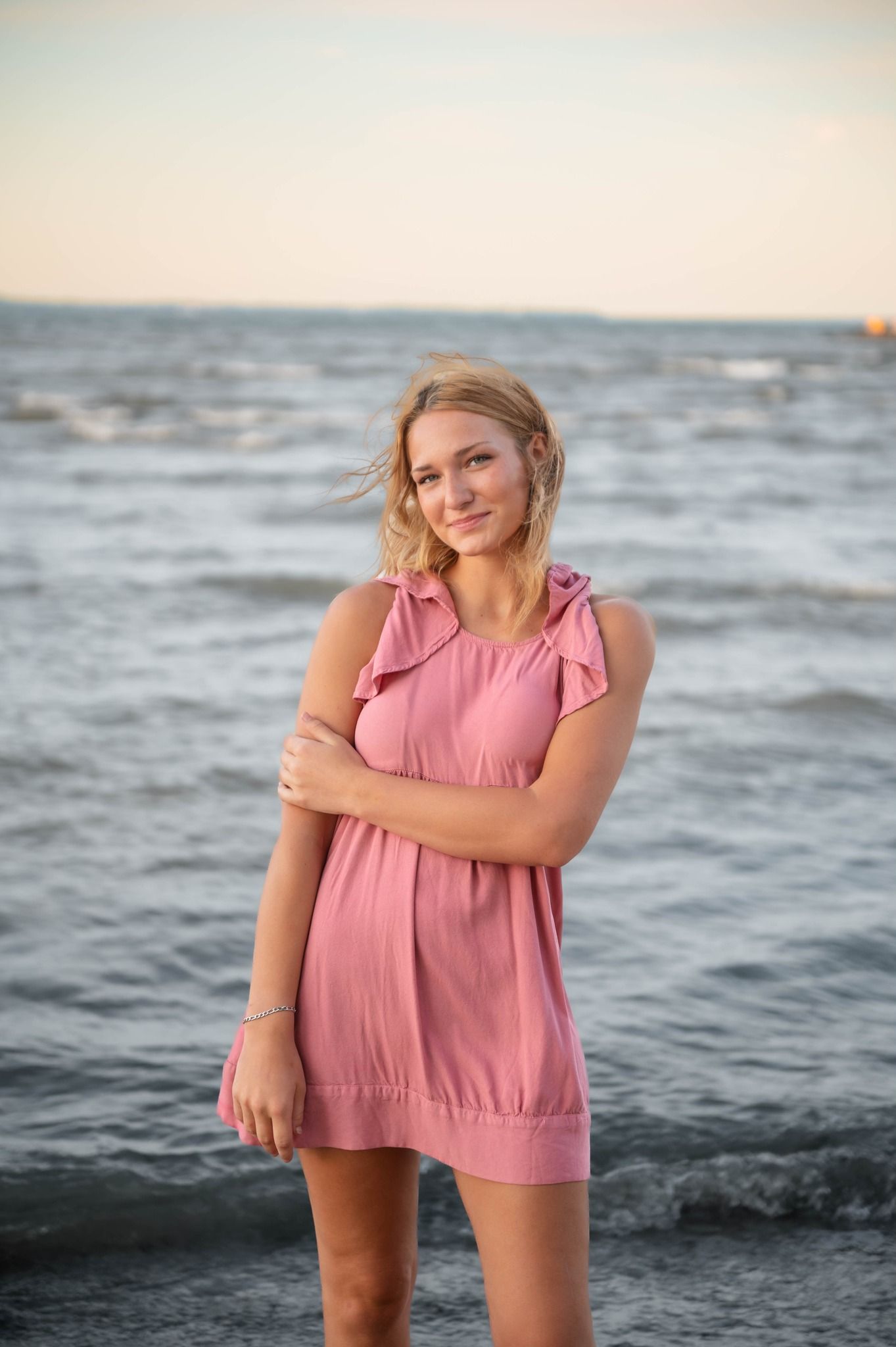 A person in a pink summer outfit poses casually by the ocean waves at dusk.