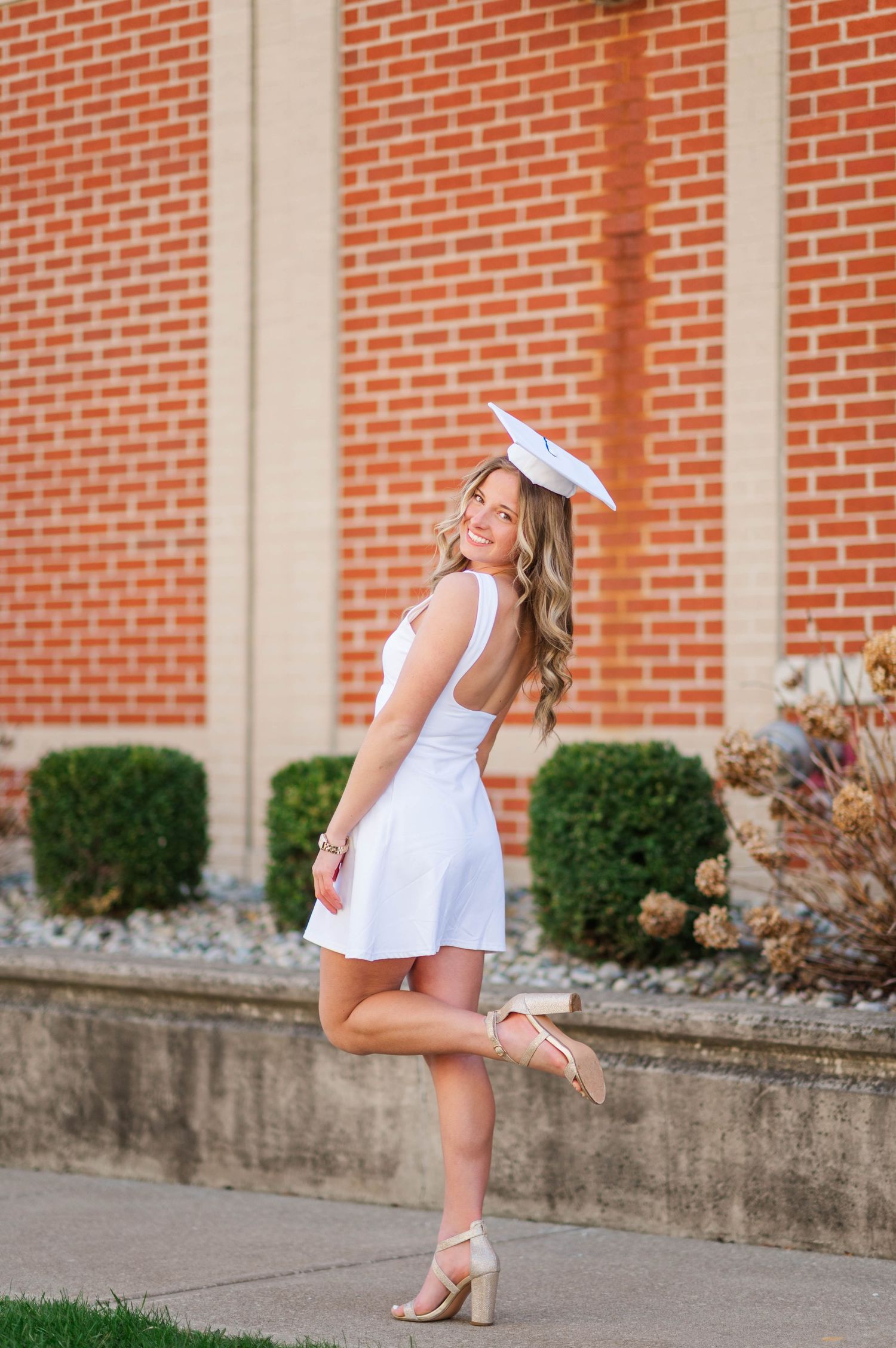 Figure in a white dress poses playfully against a brick wall background.