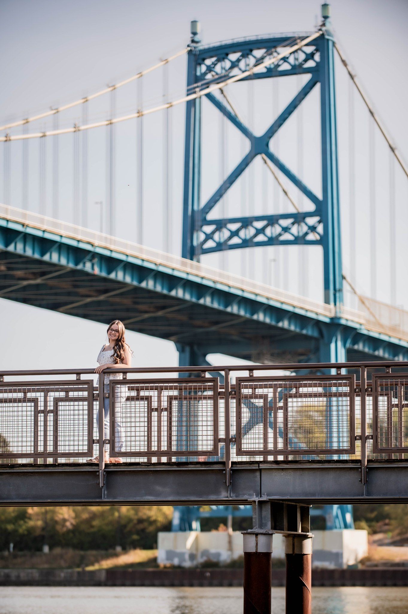 Woman poses on a waterfront railing with a large suspension bridge towering behind her on a clear day.