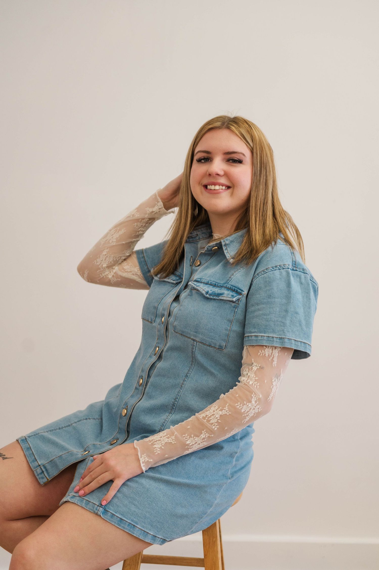 Smiling blonde woman in denim shirt dress with lace sleeves seated on stool against white background.