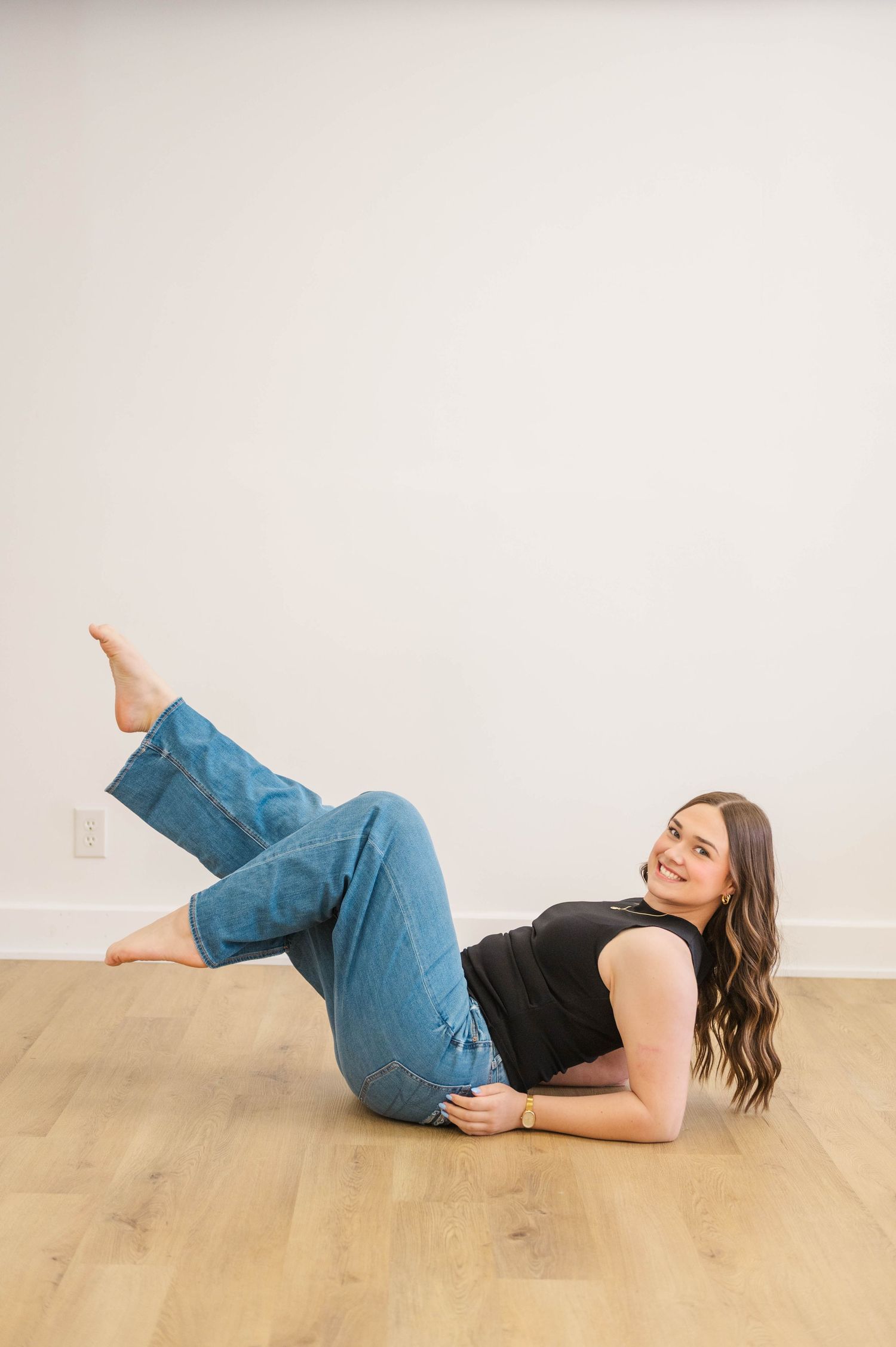Young woman with brown wavy hair lies on hardwood floor in black top and jeans, smiling over shoulder.
