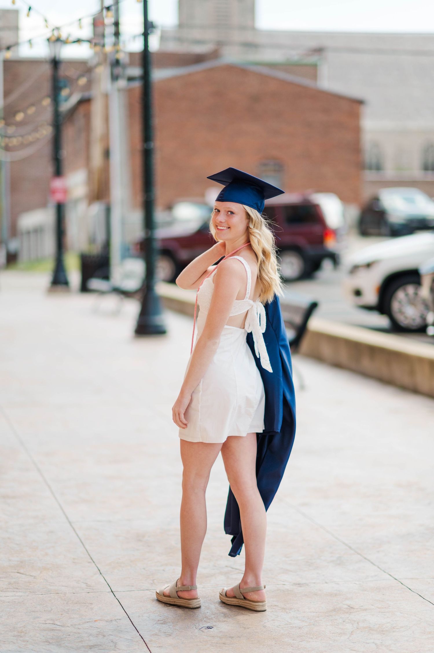 Blonde graduate in navy cap and gown smiling over shoulder on sunny urban sidewalk.
