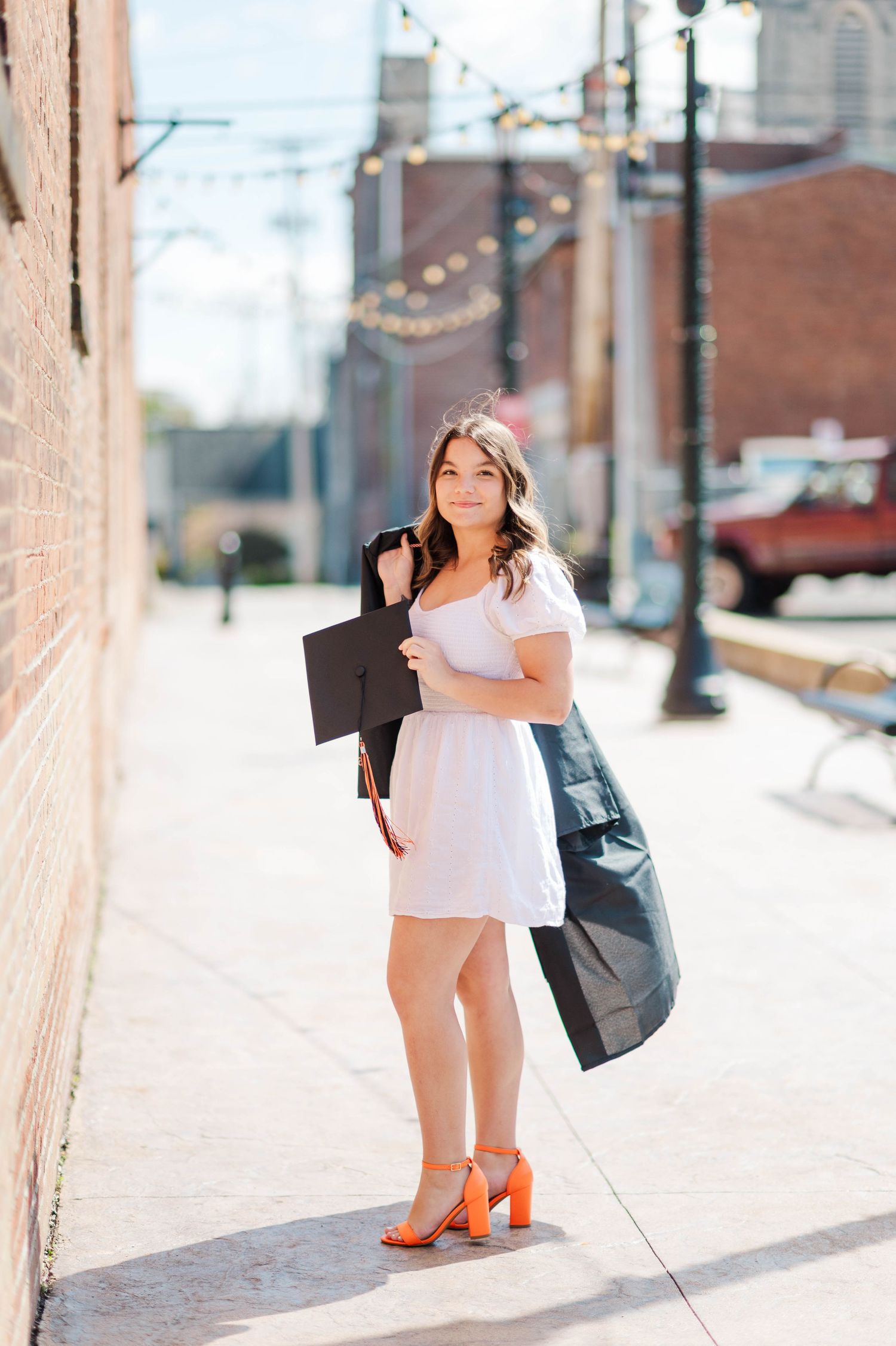 Young woman in white dress holding graduation cap and gown on urban sidewalk wearing orange heels.