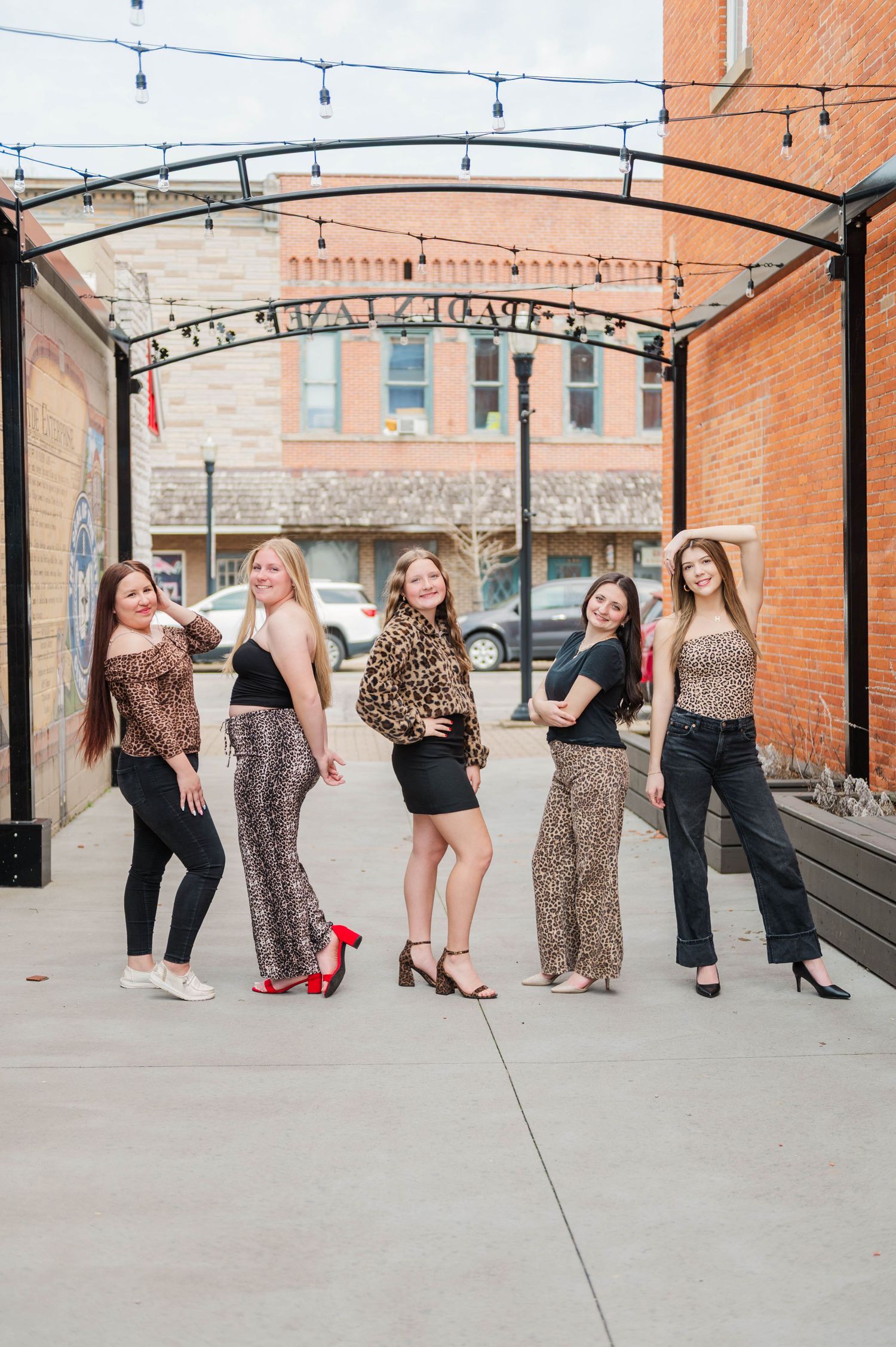 Five young women posing together under decorative arch in stylish outfits including leopard print on urban sidewalk.